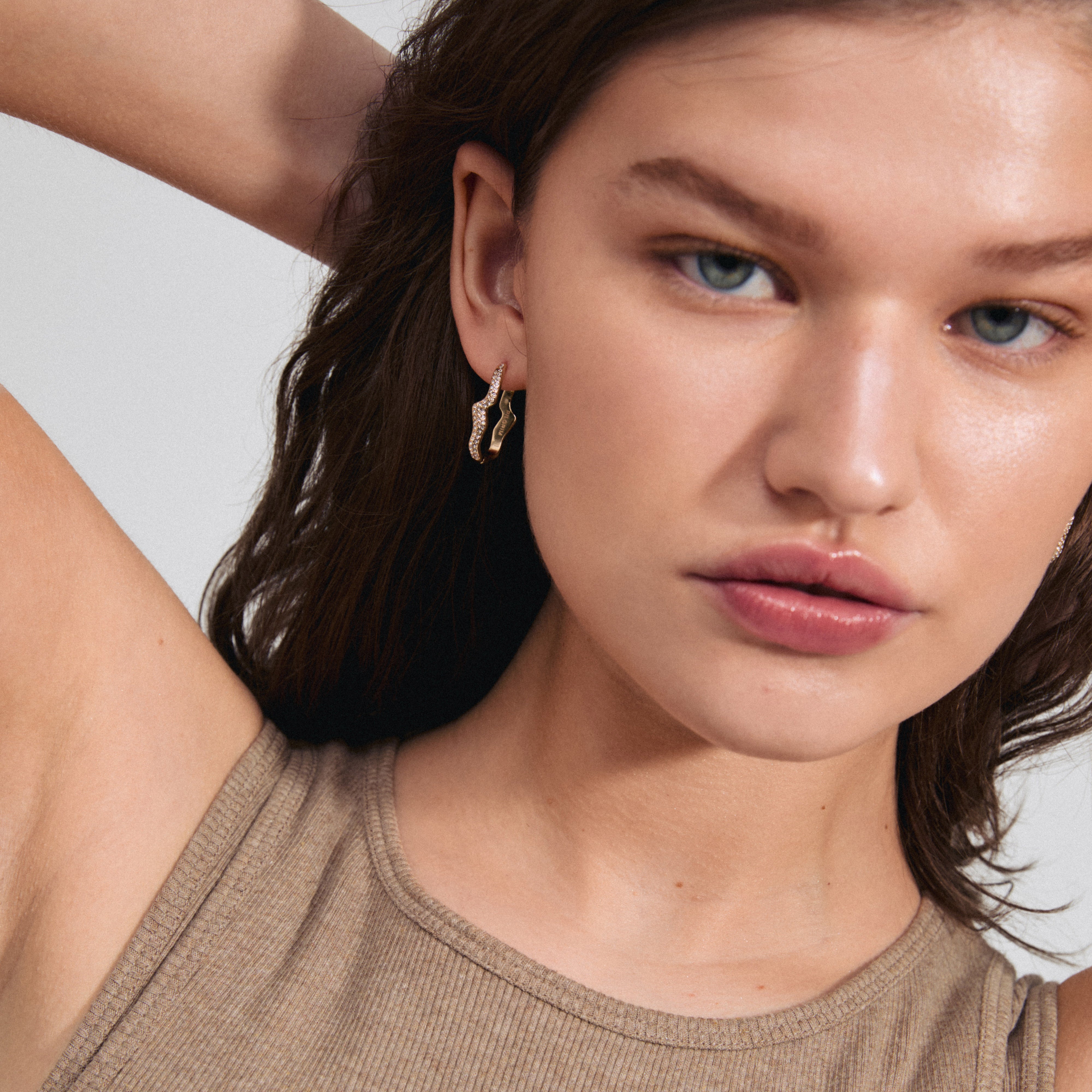 Gold-plated CONNECT crystal earrings with wavy oval hoops, featuring shiny Preciosa crystals and a click clasp. Model showcases the earrings in a styled pose.