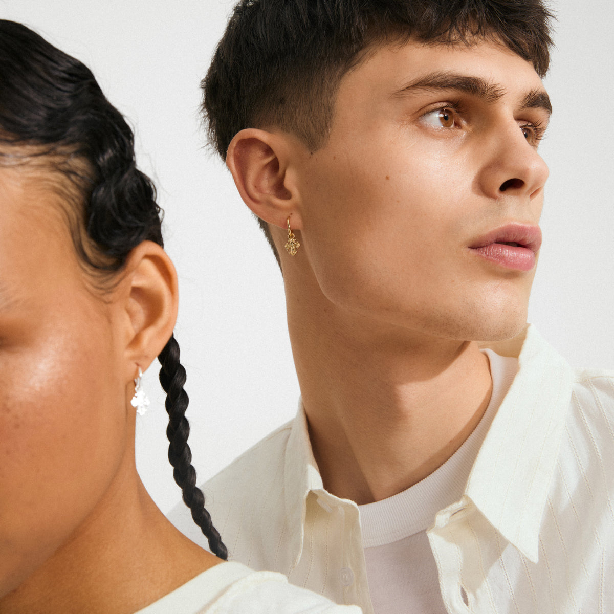 Two models showcase Pilgrim's DAGMAR huggie hoop earrings, gold-plated with floral motifs, highlighting Scandinavian craftsmanship and modern elegance in a clean, minimal style.