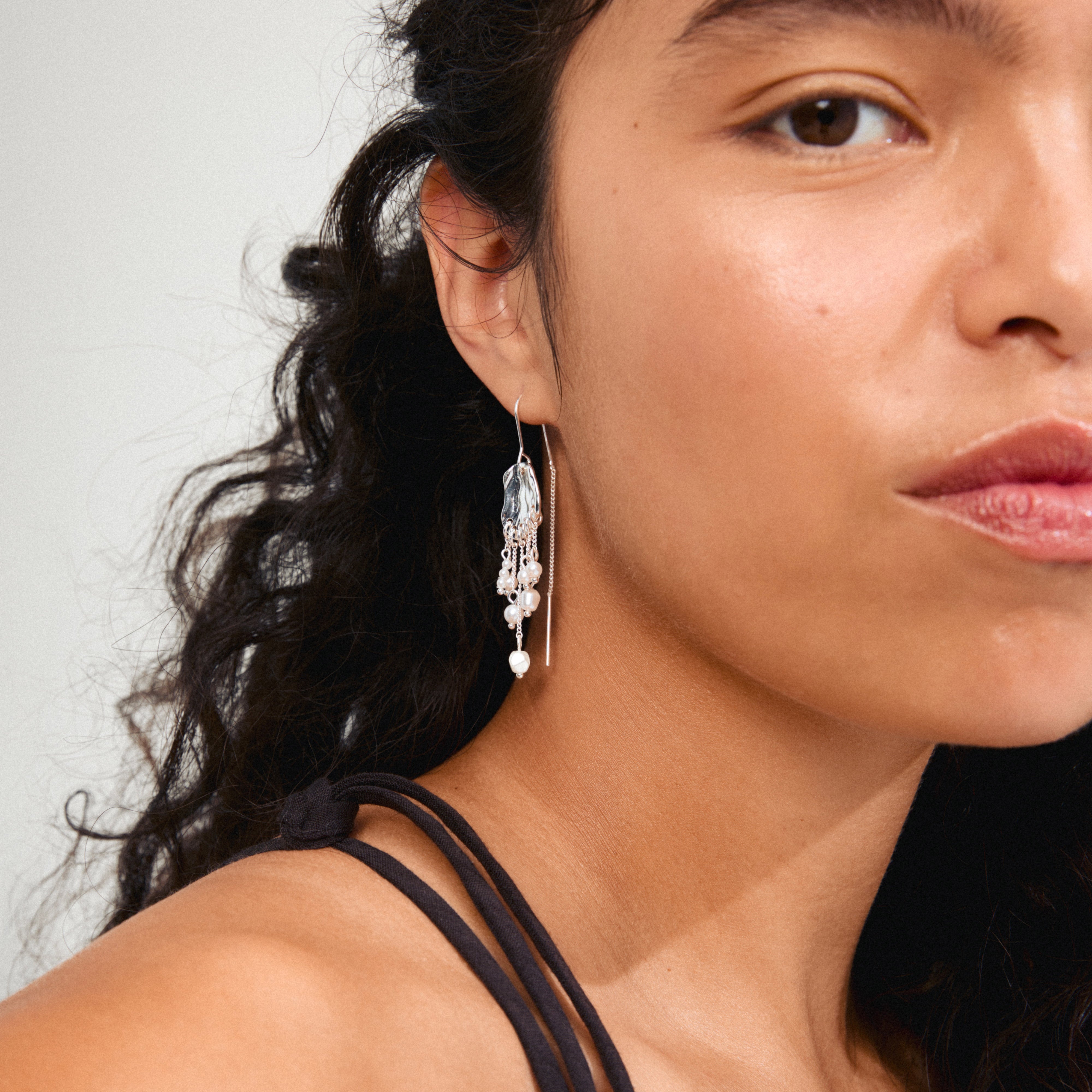 BREEZE chain earrings silver-plated, featuring asymmetrical chains with silver pendants and freshwater pearls, worn by a woman with long curly hair.