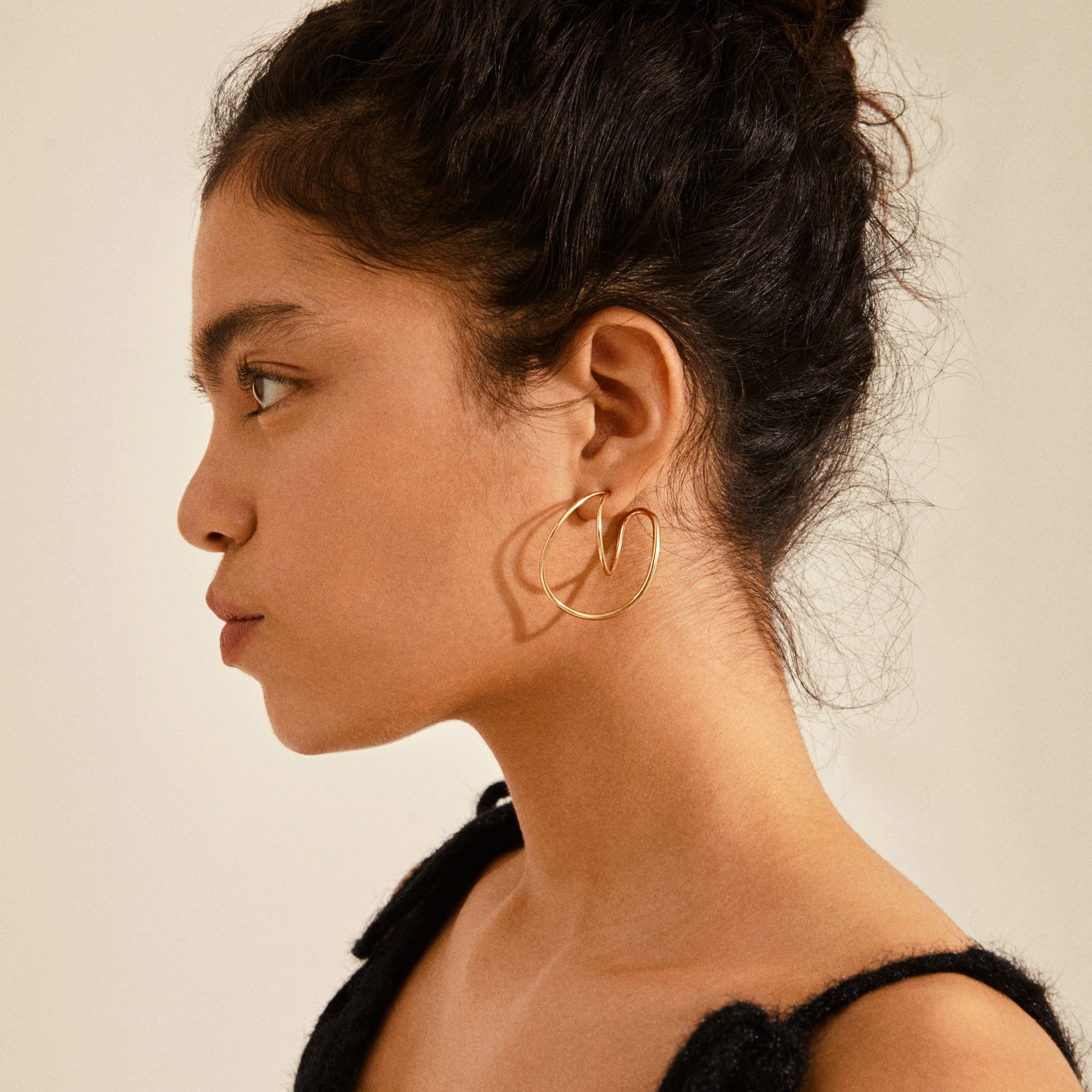Young woman wearing Pilgrim's MILLER graphic statement earrings, gold-plated, with sculptural, heart-like design, complementing her sleek black top.