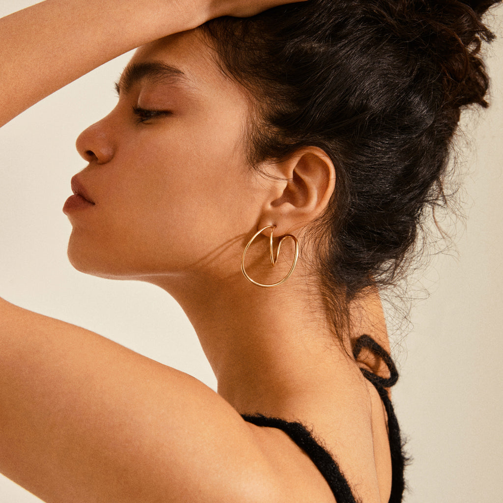 Close-up of a young woman wearing the Pilgrim MILLER gold-plated earring, showcasing its sculptural design with a heart-like curve at the base.
