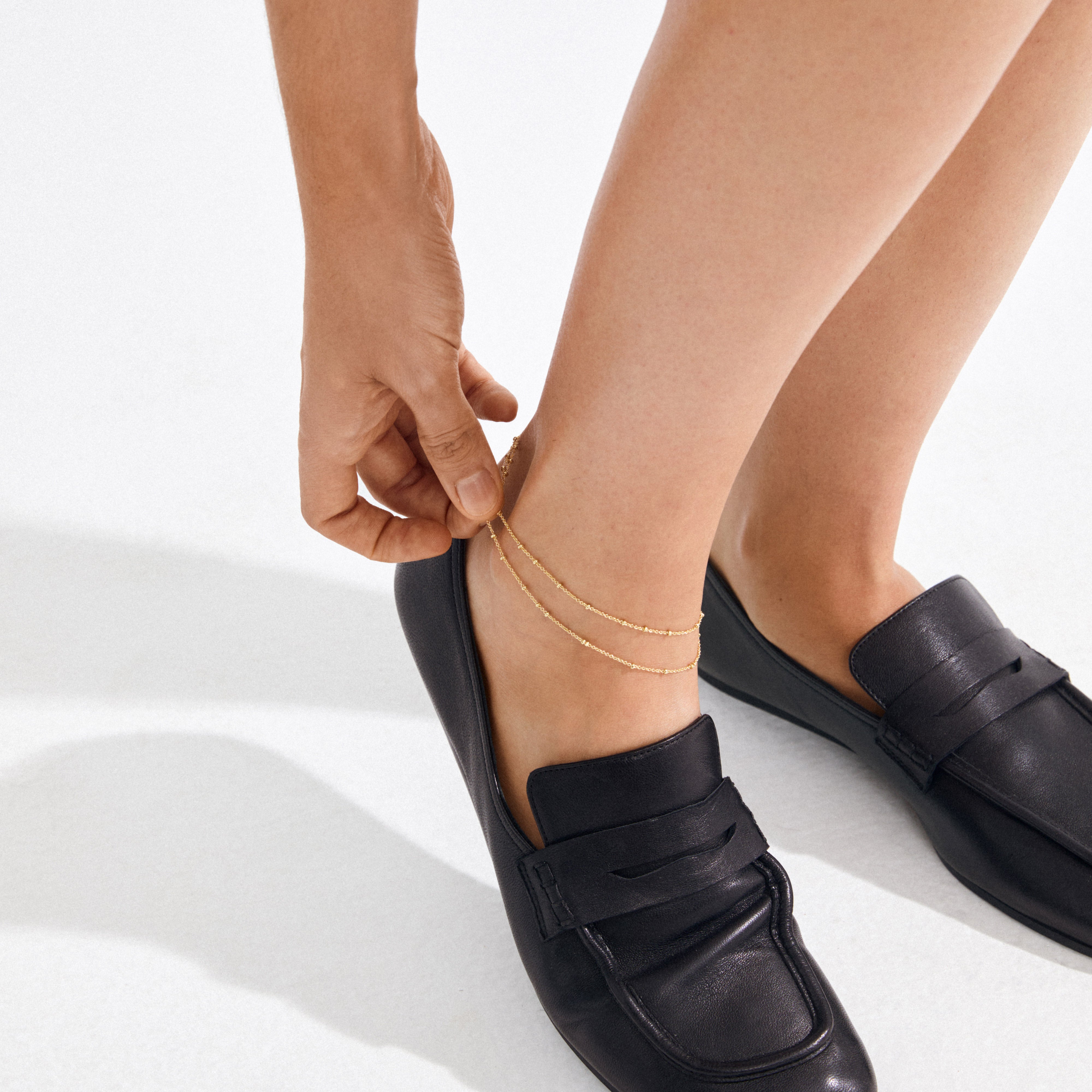 A person adjusts the ELKA ankle chain 2-in-1 gold-plated, showcasing its sleek double strands and tiny pendants over black leather loafers.
