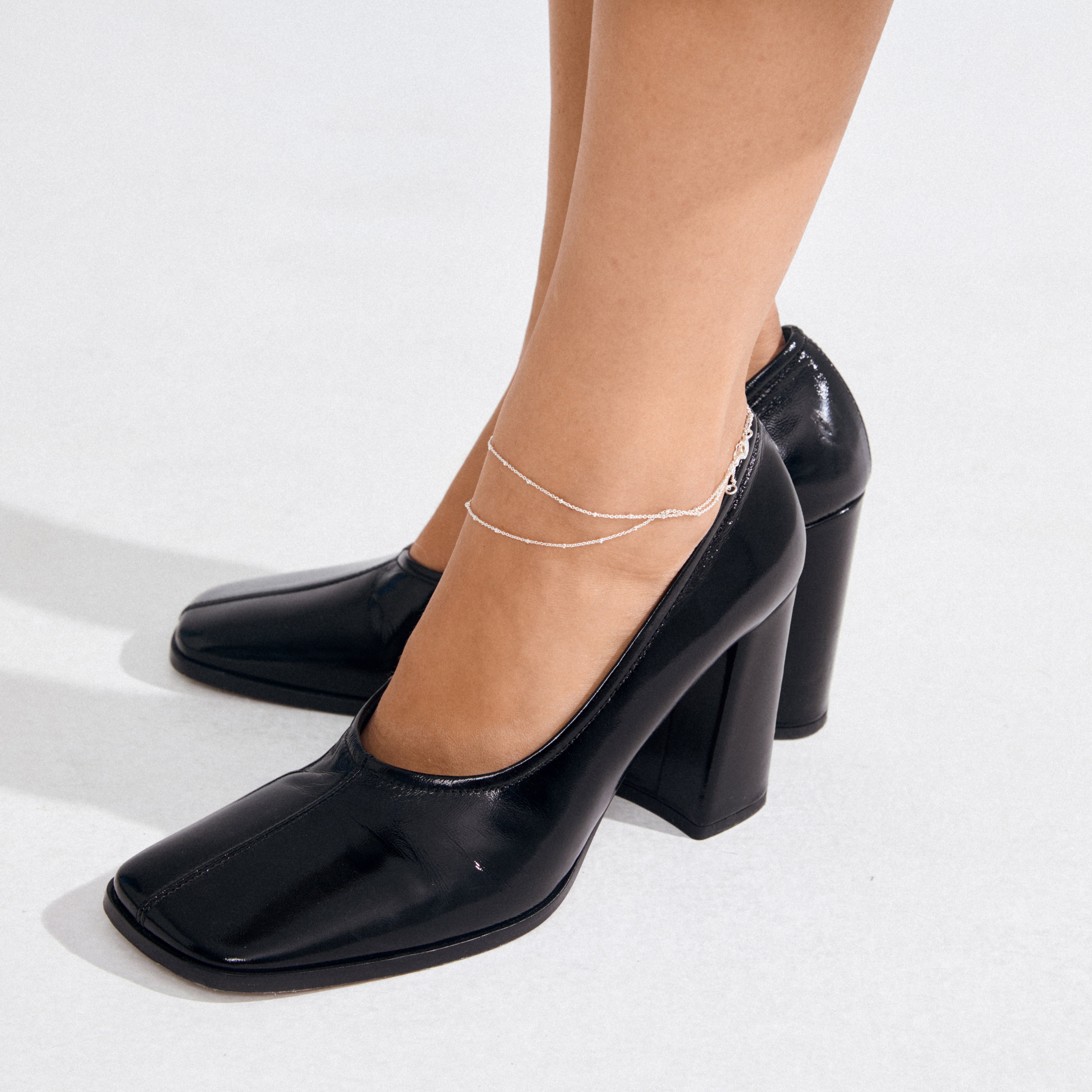 Model wearing glossy black leather pumps with the ELKA 2-in-1 silver-plated anklet, featuring delicate double-strand chains and circular pendants, showcasing Pilgrim's chic style.