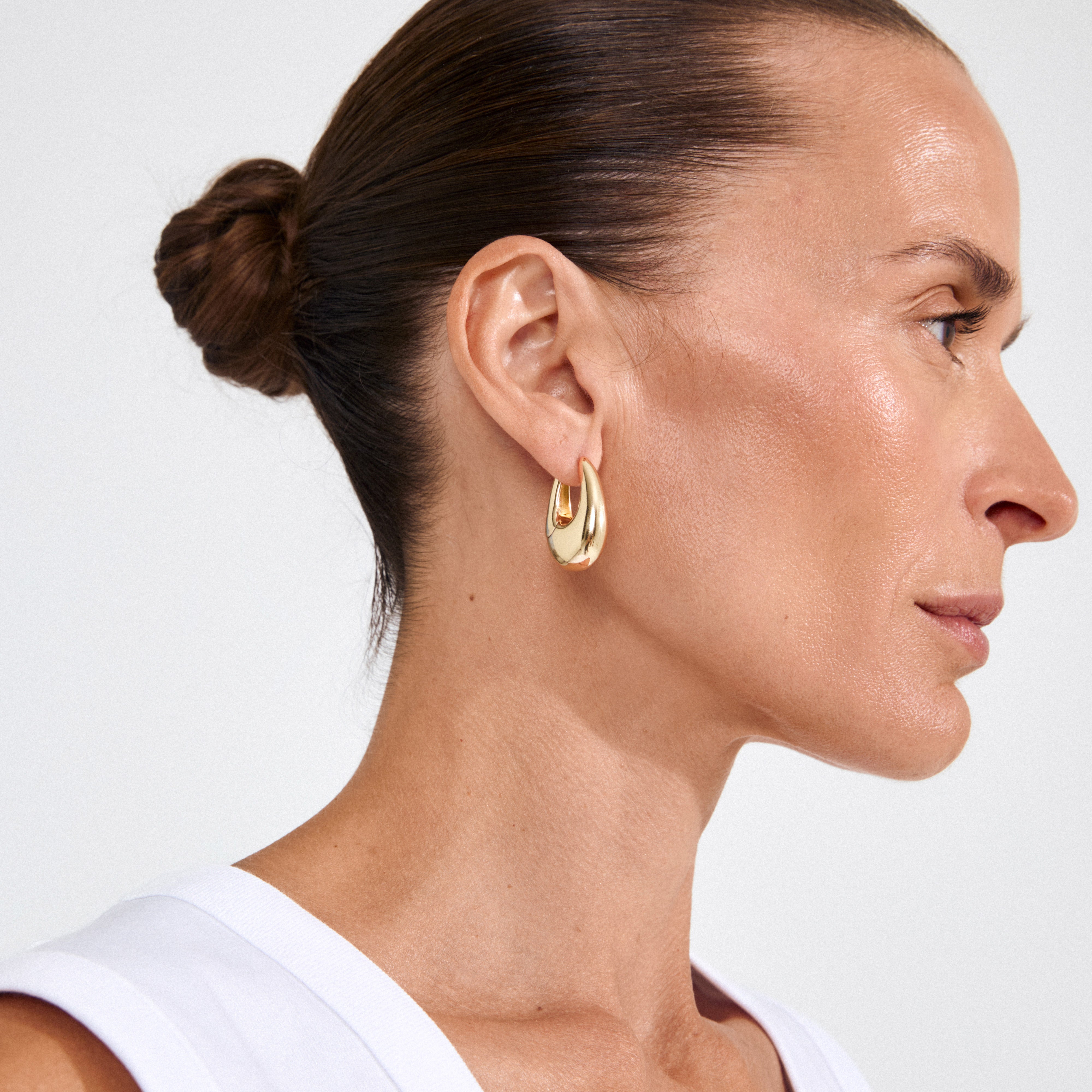 Side profile of a woman wearing Pilgrim's AUTUMN chunky retro hoop earrings, gold-plated, showcasing their bold, teardrop design against her minimalist outfit.
