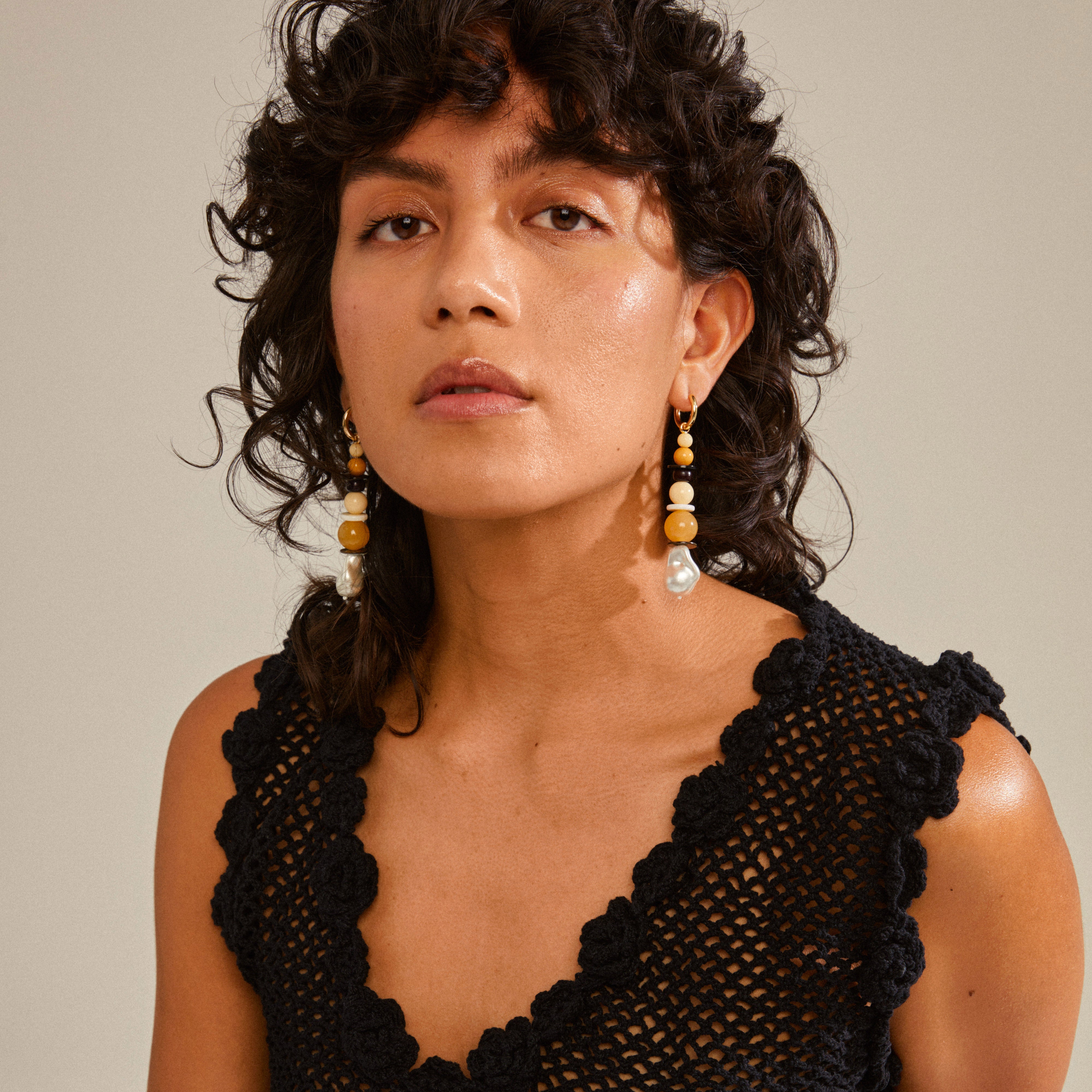 Woman with curly hair wearing NAILA earrings; gold-plated, multi-colored beaded hoops by Pilgrim, enhancing her black dress ensemble.