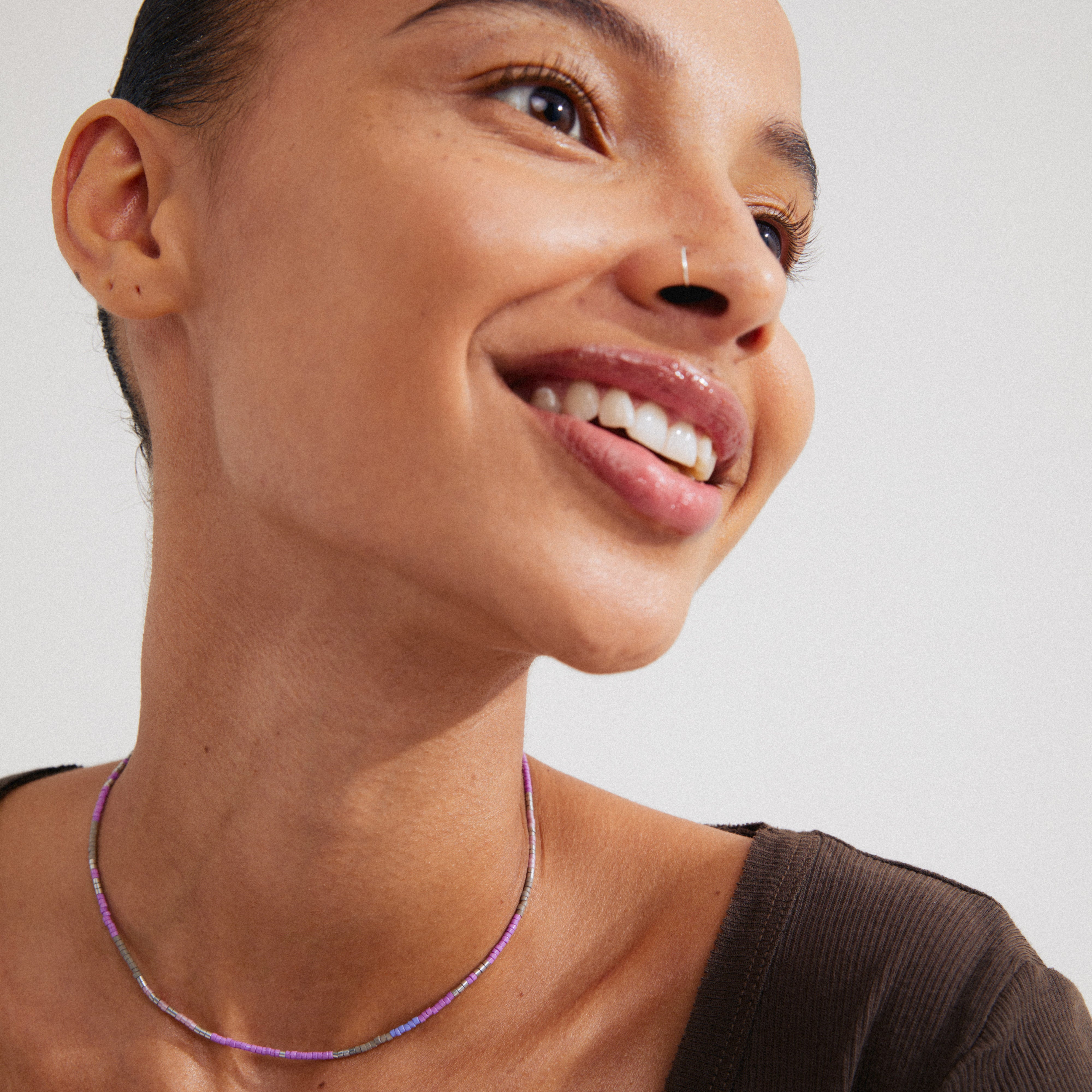 Woman smiling, wearing Pilgrim's ALISON necklace featuring purple and grey glass beads, providing a boho-inspired, layered look. Perfect for a festival-ready style.