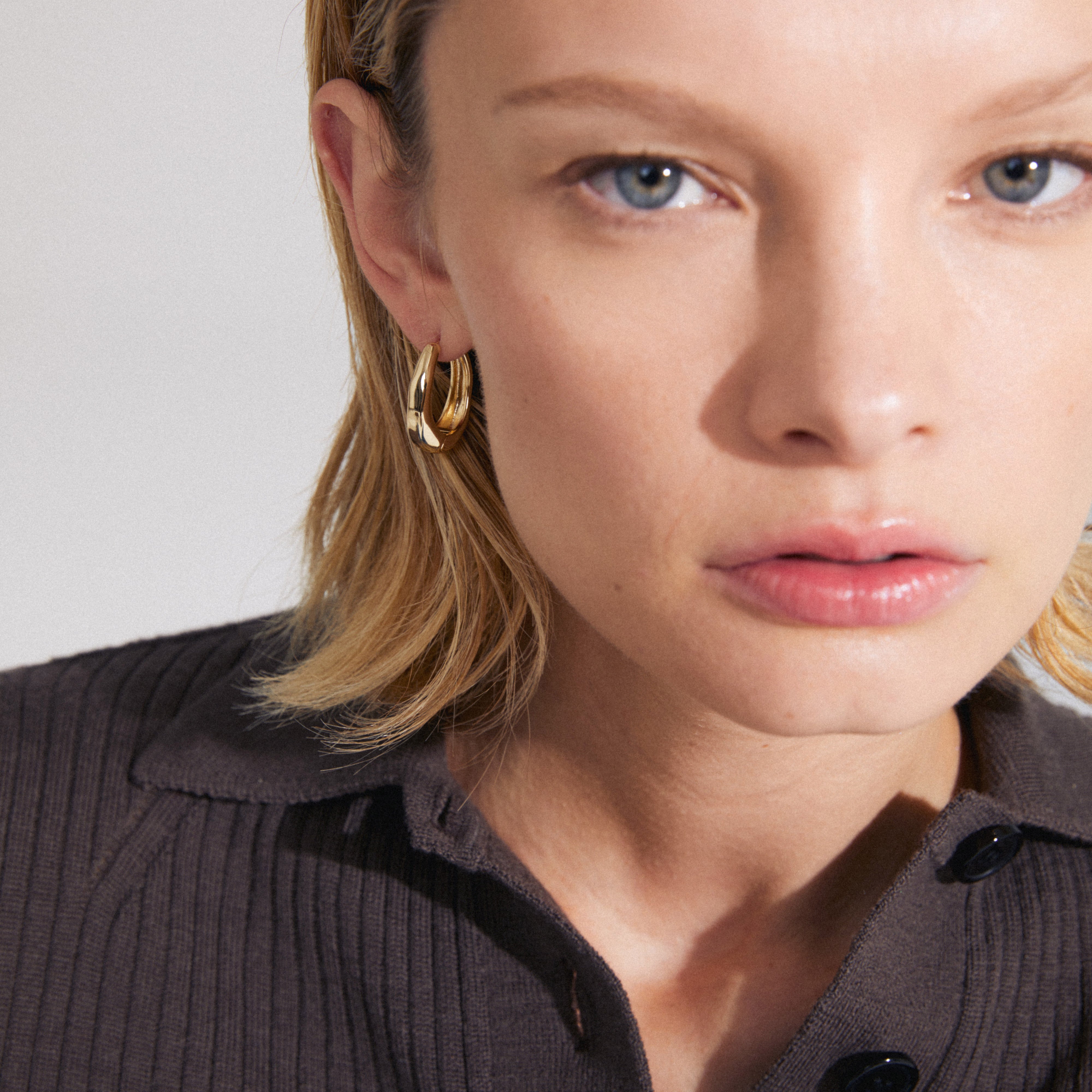 Close-up of a woman wearing ASHER gold-plated earrings, showcasing their chunky oval design and easy click clasp. Perfect blend of trendy and timeless style.