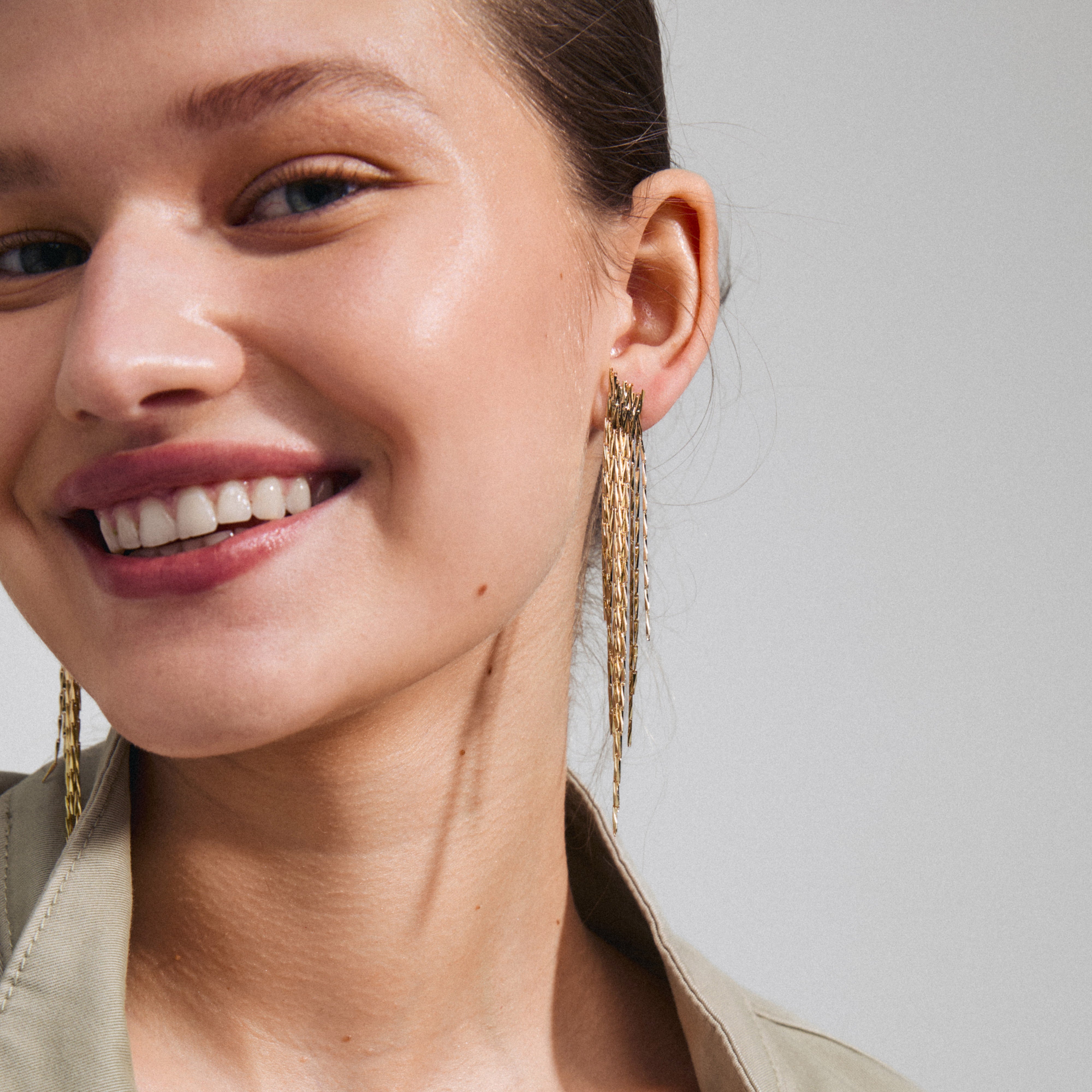 Woman smiling while showcasing ZENOBIA gold-plated earrings with layered chains and twisted melon seed links, designed to sparkle and perfect for parties.