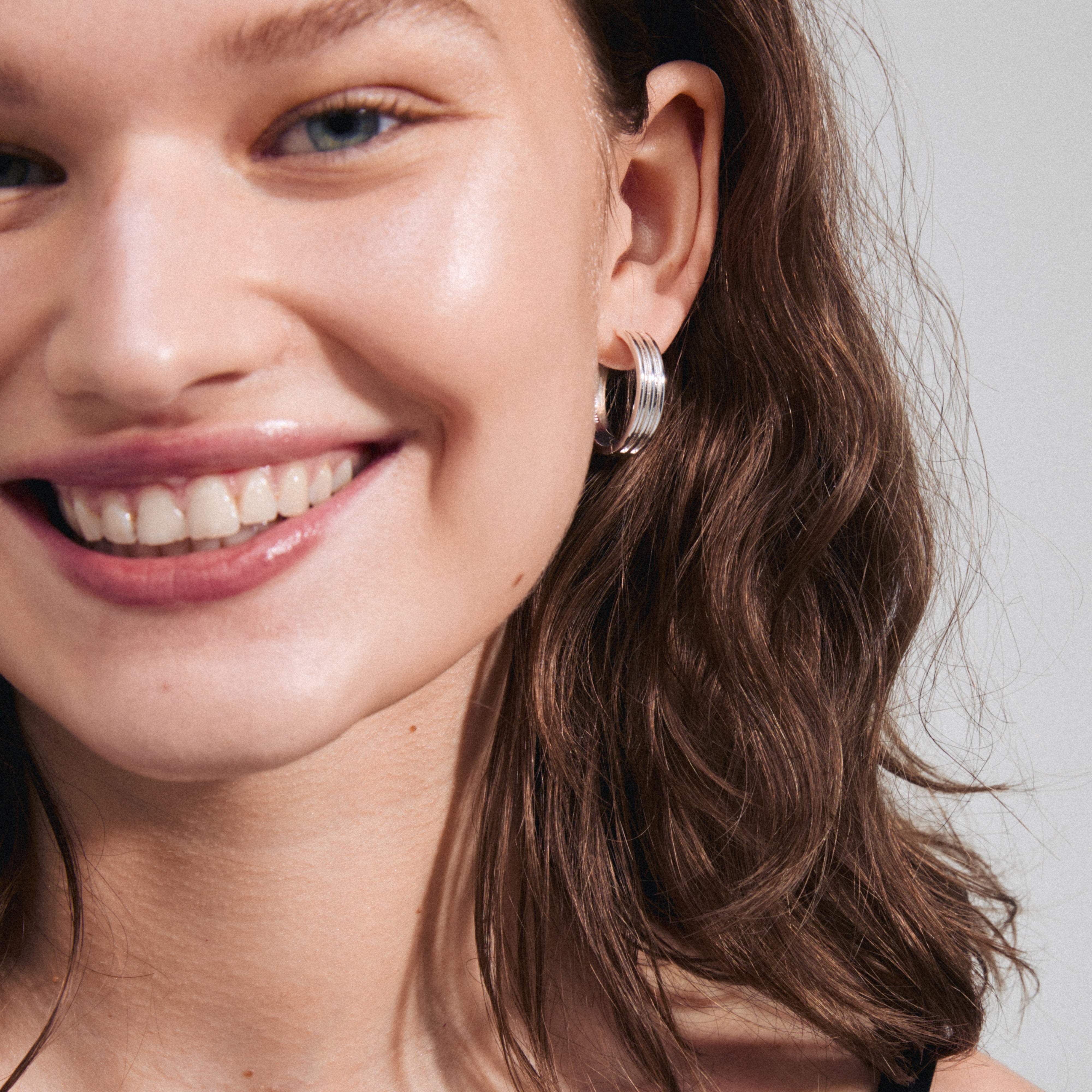 Woman smiling, showcasing BENNETT hoop earrings silver-plated from Pilgrim, highlighting their classic, wide design and urban-inspired simplicity.