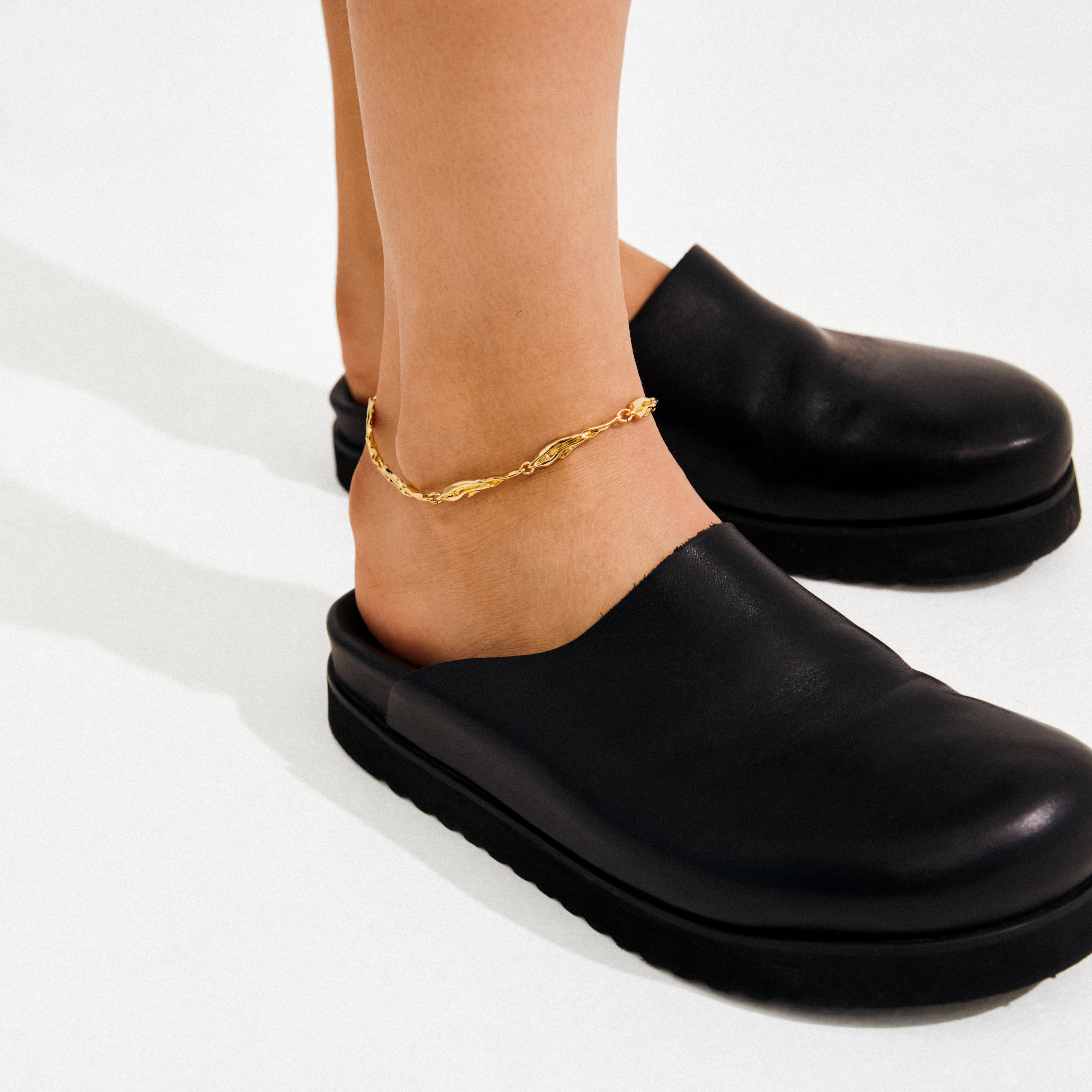 Person's lower legs wearing ARIF gold-plated anklet with organic, elongated links, paired with black leather mules. A stylish summer accessory from Pilgrim.