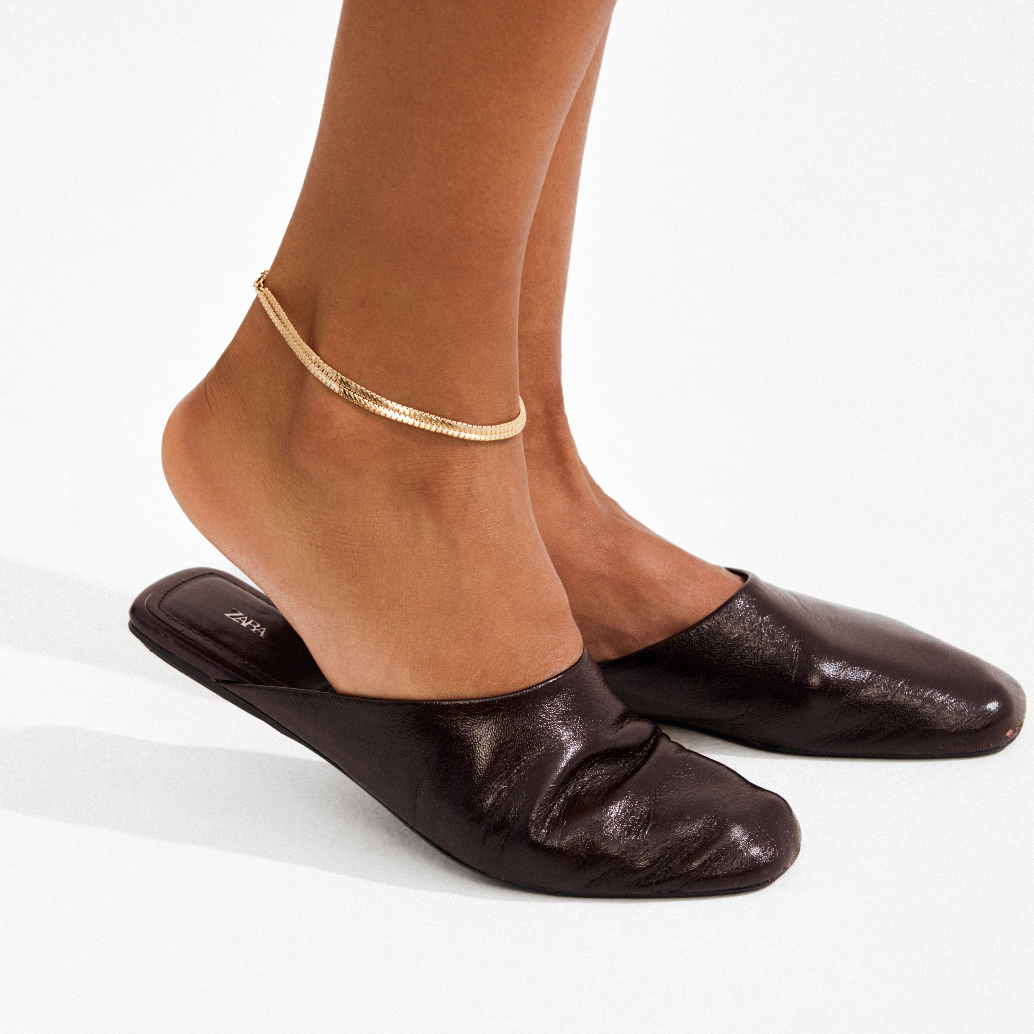 JELONA ankle chain gold-plated with dual snake chains shines on ankle, paired with brown leather mules, highlighting Pilgrim's modern Scandinavian style.