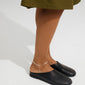 The JOANNA silver-plated anklet elegantly adorns an ankle, featuring a herringbone chain with a charm, paired with black leather mules for a chic summer look.