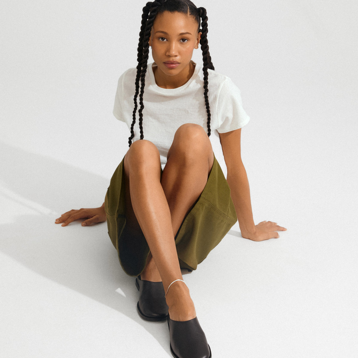 Young person wearing a silver-plated JOANNA anklet, styled with minimalist black slip-ons and an olive skirt, highlighting its elegant, light-reflecting herringbone chain design.
