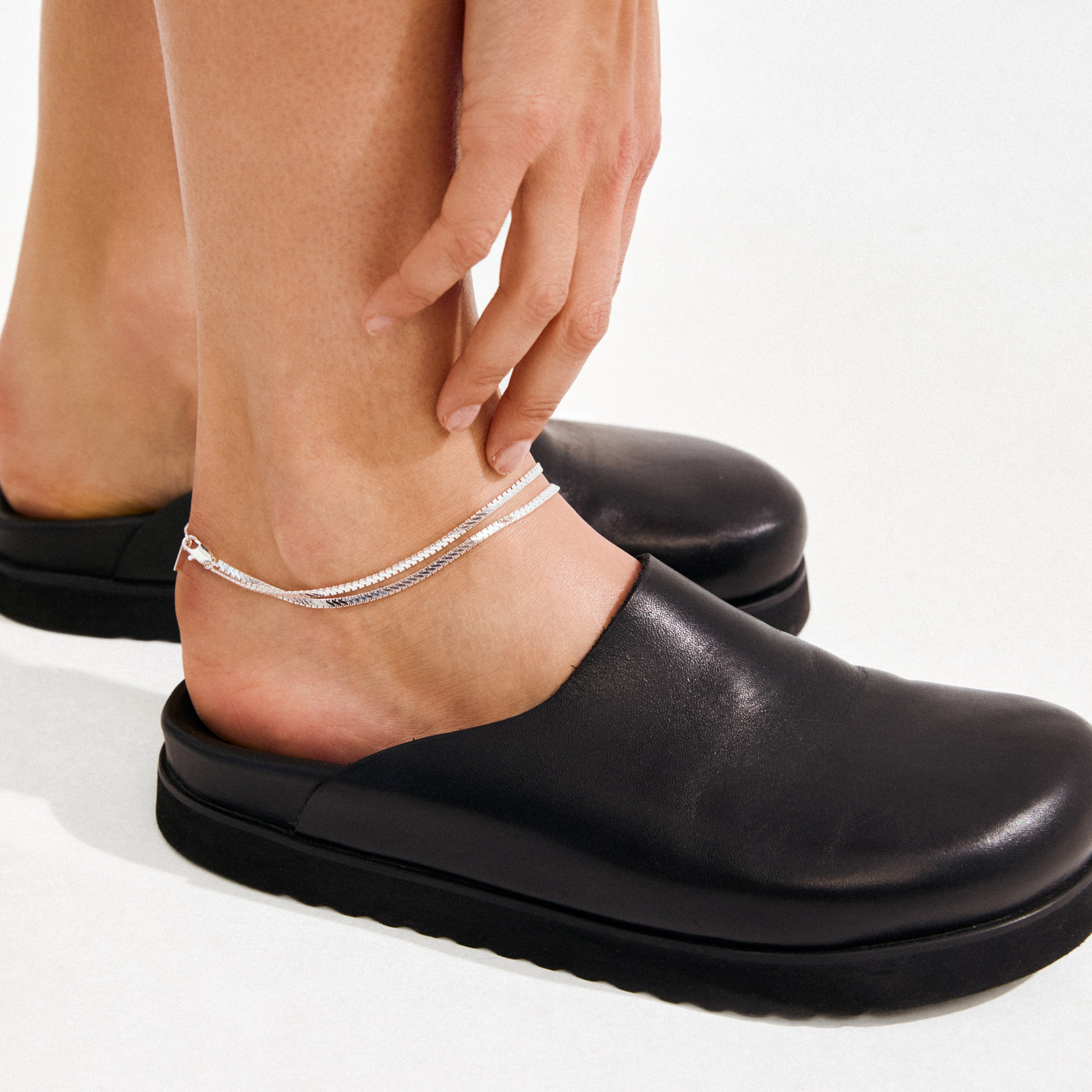 JELONA ankle chain silver-plated: Double-strand herringbone chains catch light, elegantly layered on a bare ankle, paired with a black leather mule. Perfect for summer style.