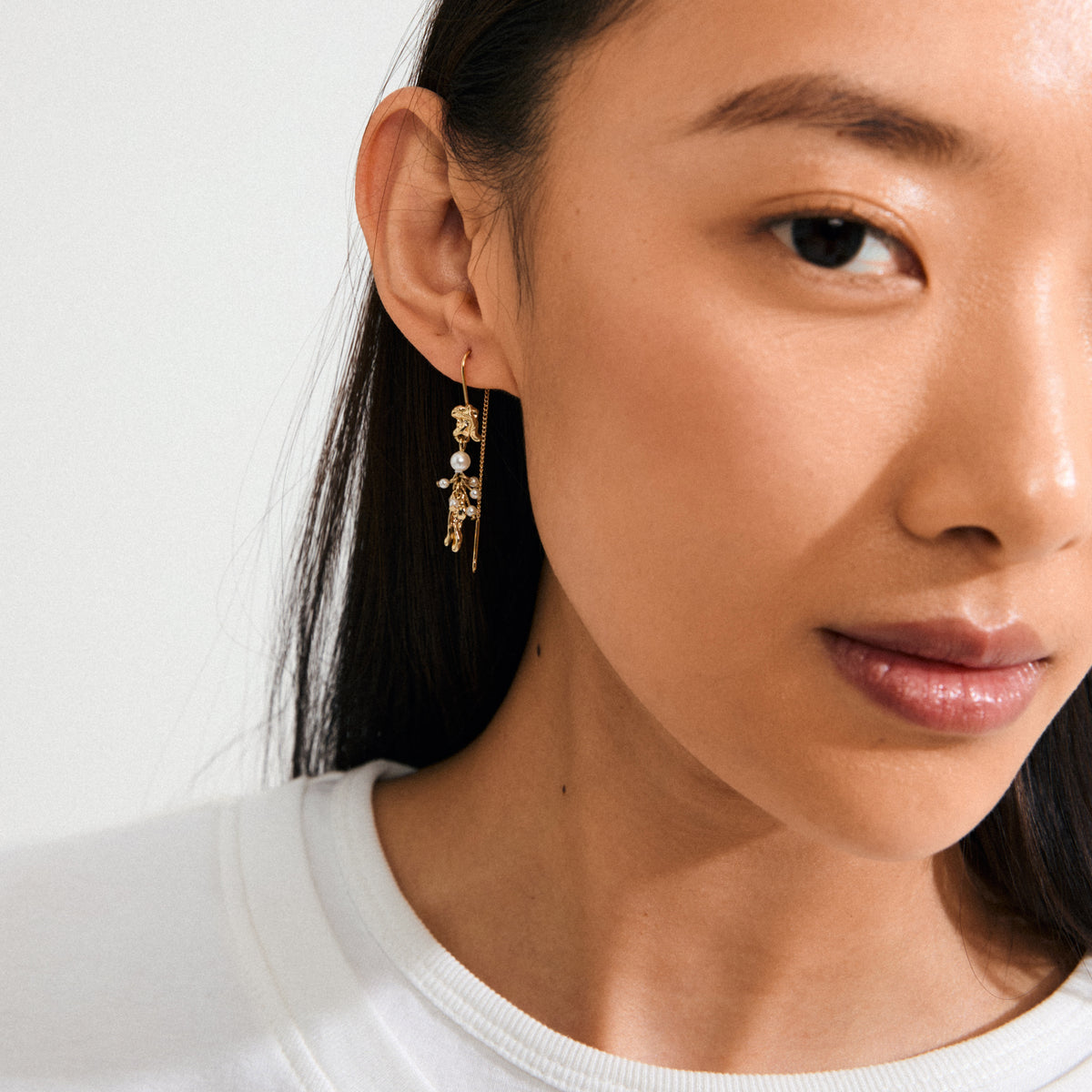 Young woman wearing MILO chain earrings gold-plated by Pilgrim, featuring pearls and charms, highlighting a whimsical, adventurous touch against her smooth skin and dark hair.