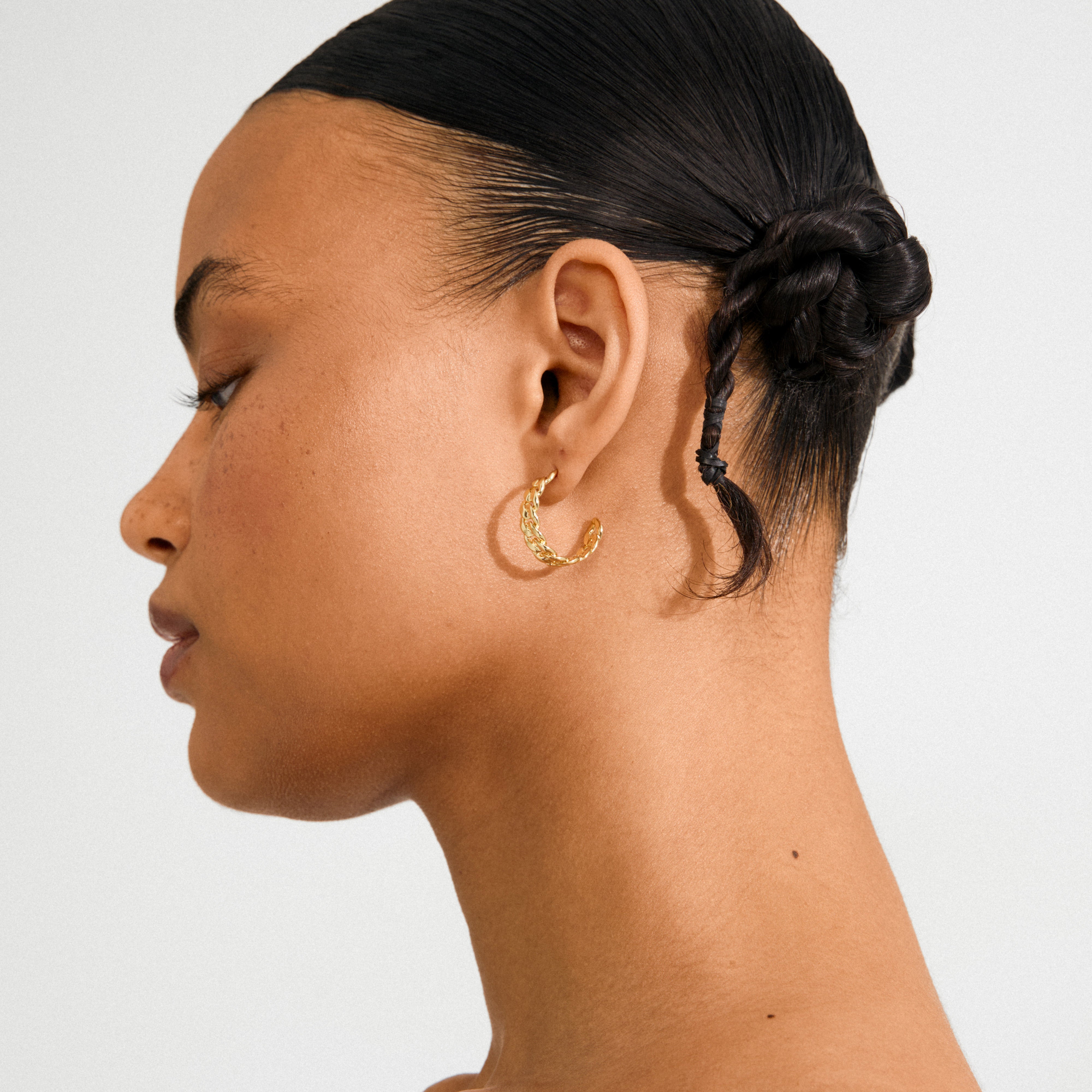 Close-up of a person wearing Pilgrim's MAYER earrings gold-plated, featuring a textured curb chain hoop, showcasing edgy elegance with a sleek braided hairstyle.