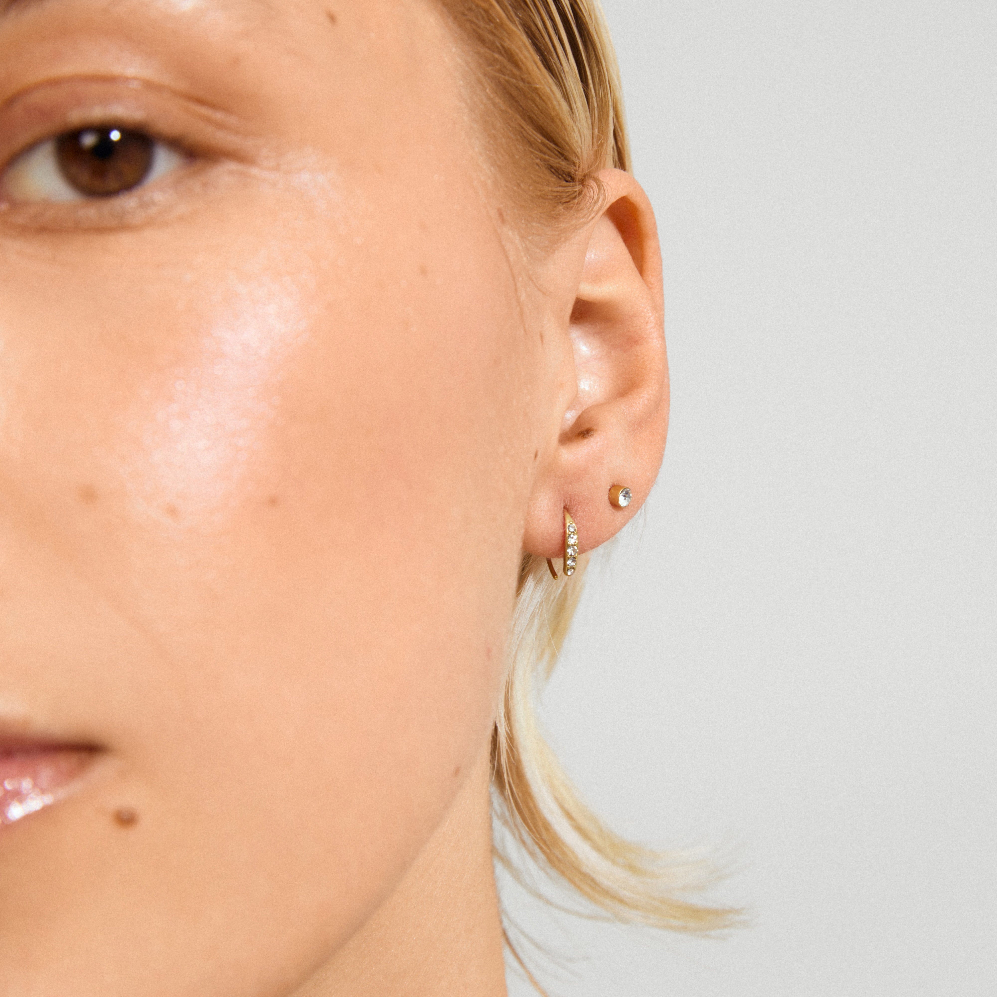 Close-up of GIONNI earrings 2-in-1 set, featuring a gold-plated stud with crystals and a gem-set hoop on a light-skinned person’s ear.