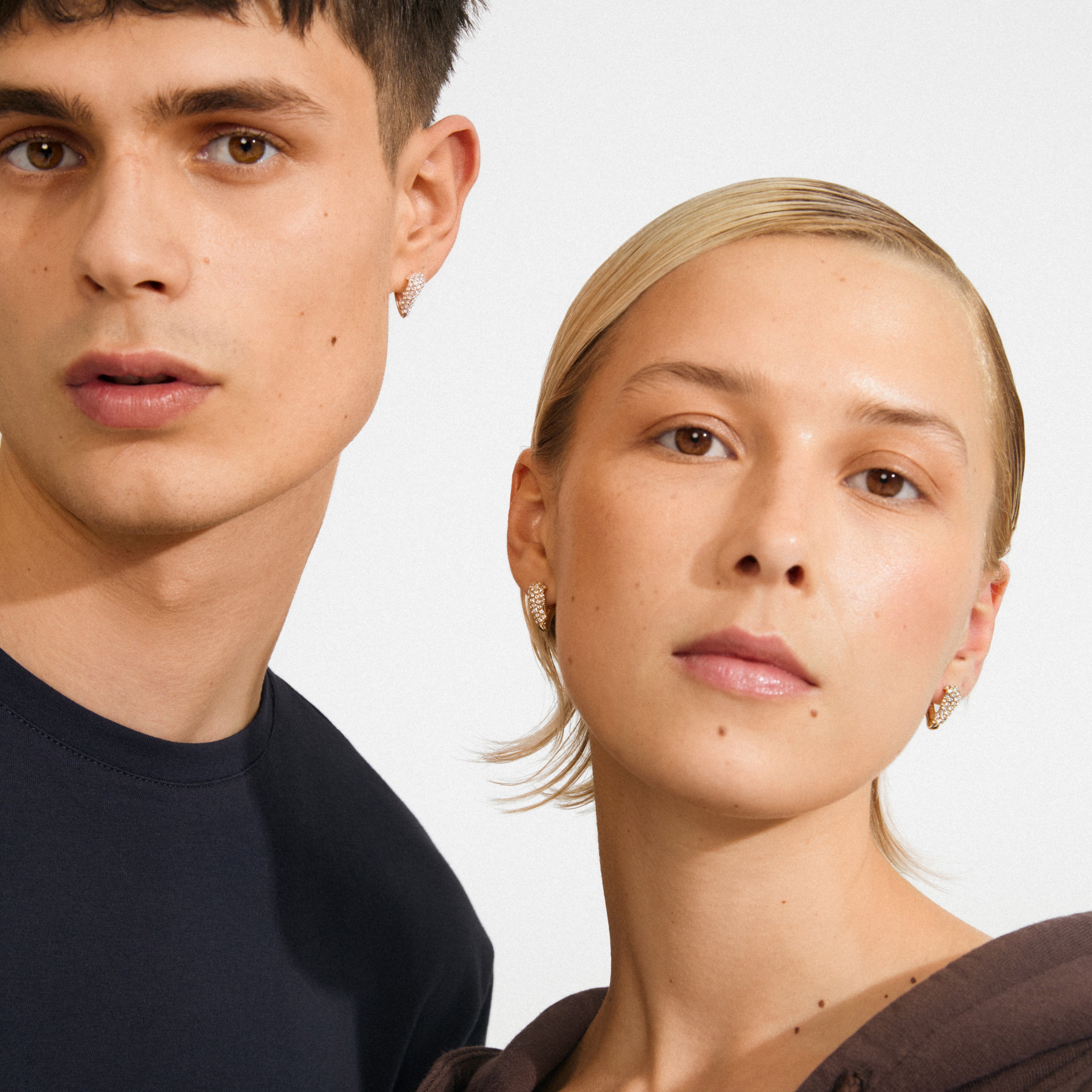 Two young adults model Pilgrim's SOUL earrings, gold-plated with pavé-style crystals, forming a heart shape, symbolizing love and connection.