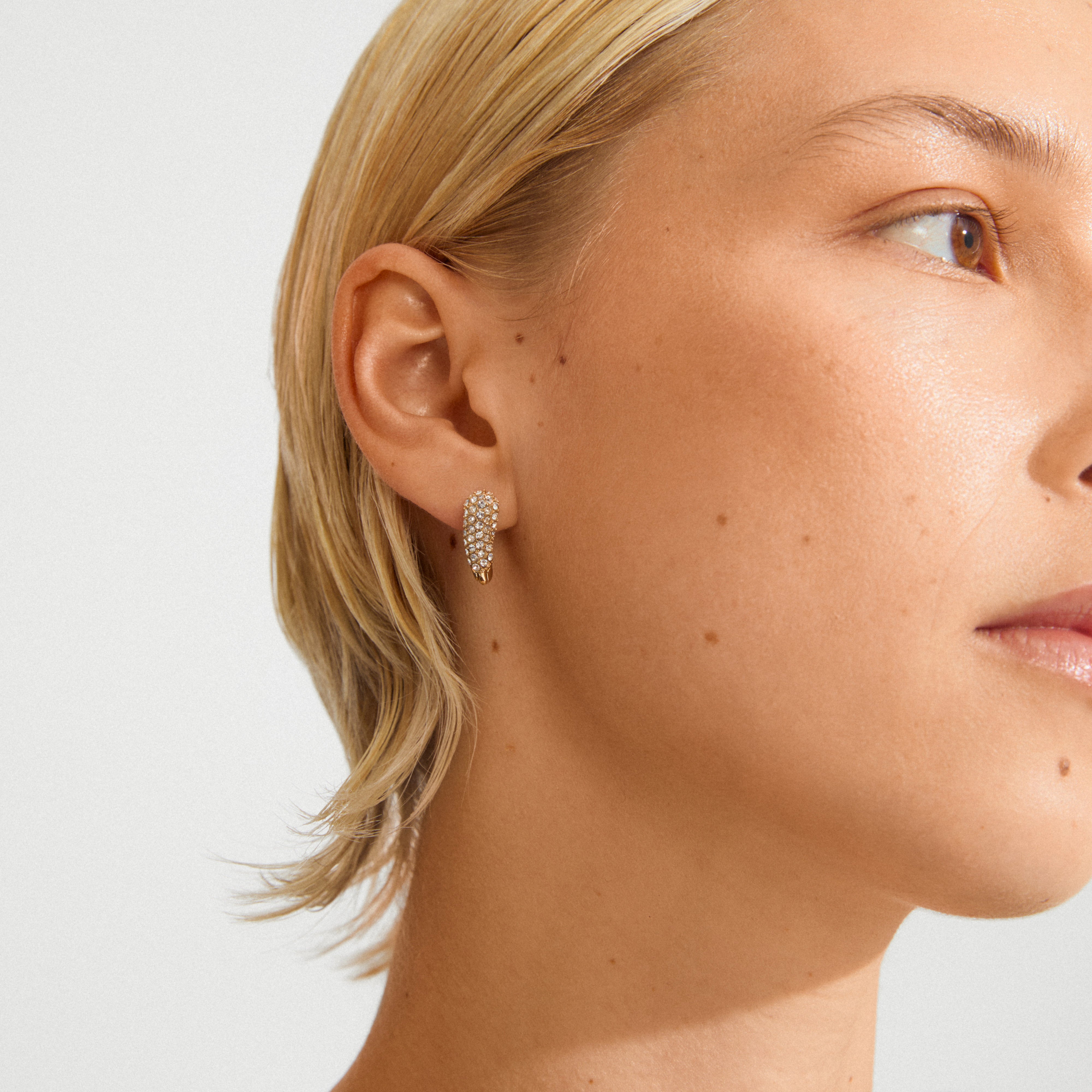 Close-up of a light-skinned person wearing Pilgrim's gold-plated SOUL earrings, featuring a delicate heart shape with clear gemstones, symbolizing love and connection.