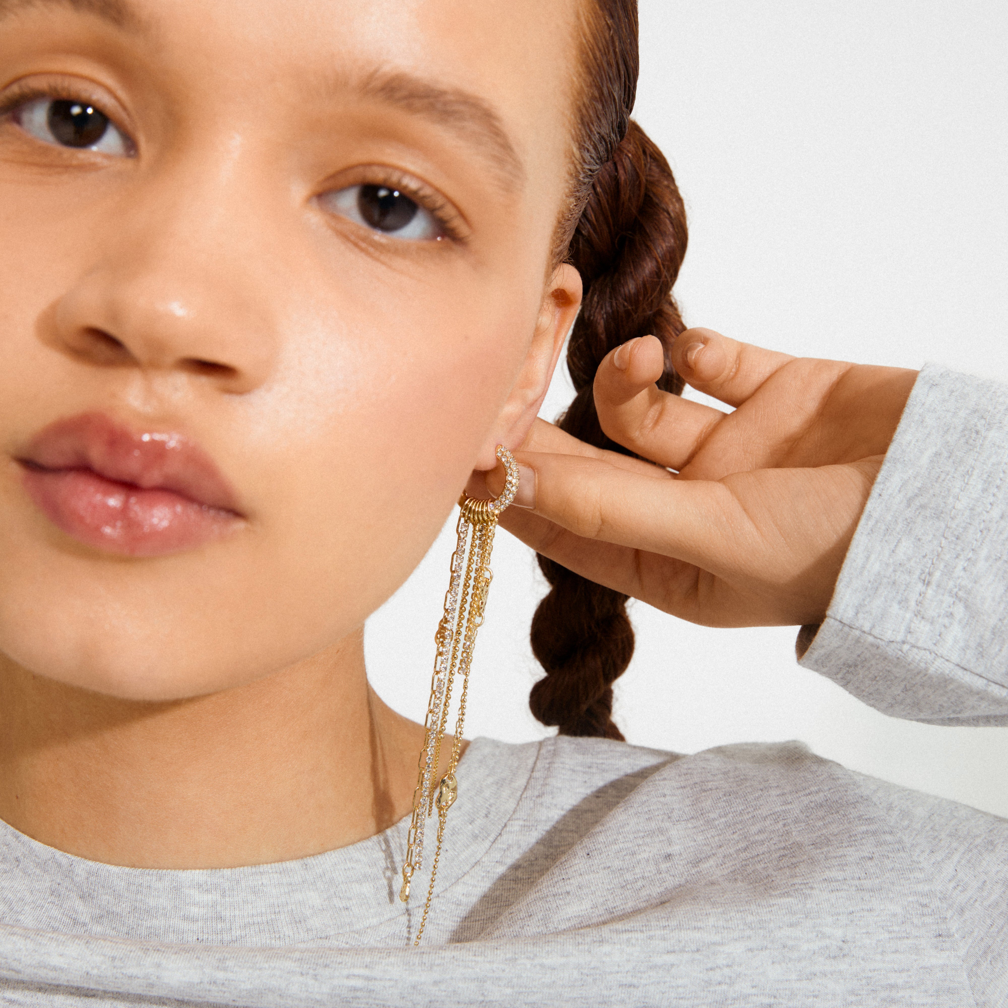Person wearing gold-plated DRUE earrings with tiny stones and dangling chains, gently touching the earring, highlighting its intricate design and luxurious detail.