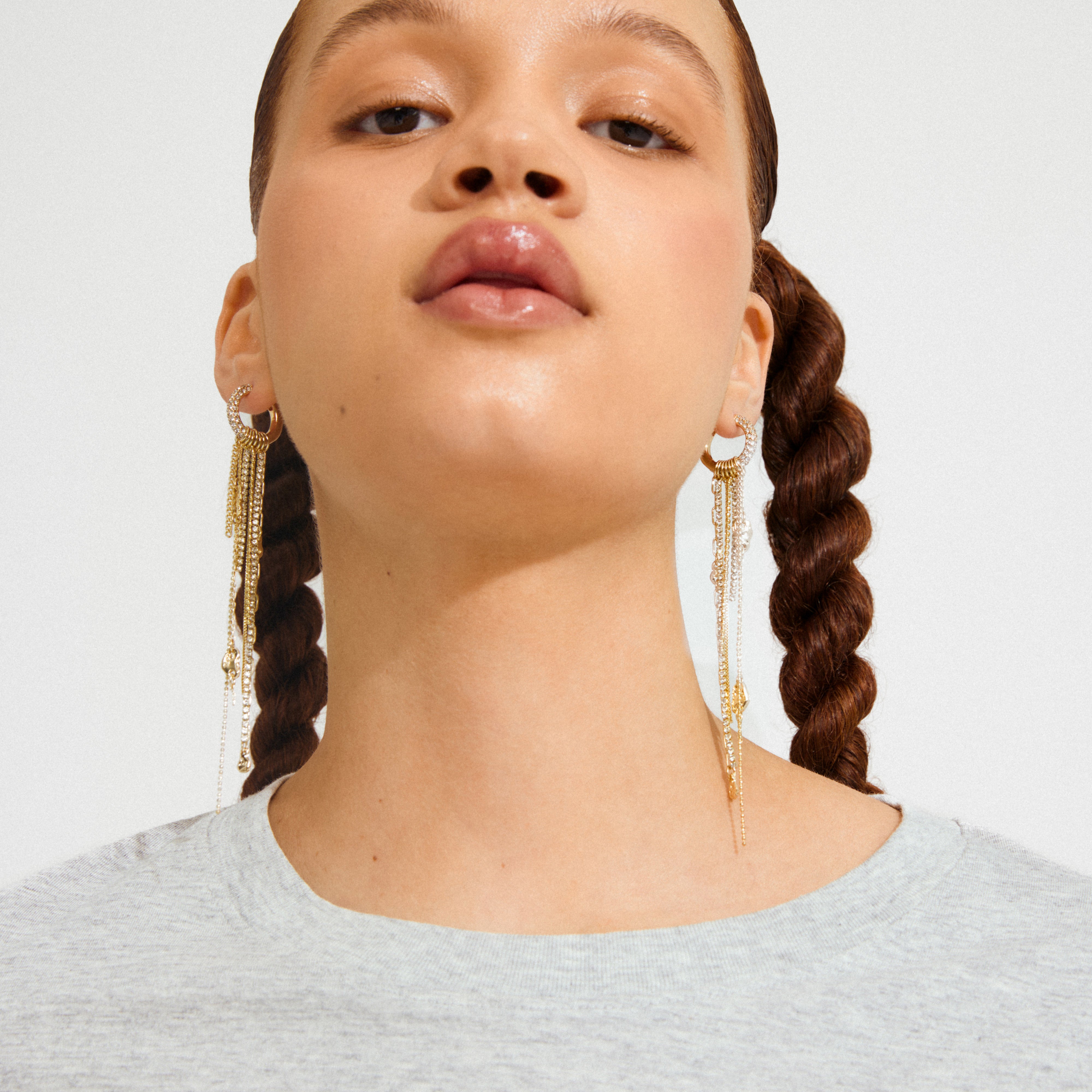 Young person wearing Pilgrim's DRUE gold-plated earrings with chain tassels, styled with braided hair and a light gray top, highlighting a chic bohemian vibe.