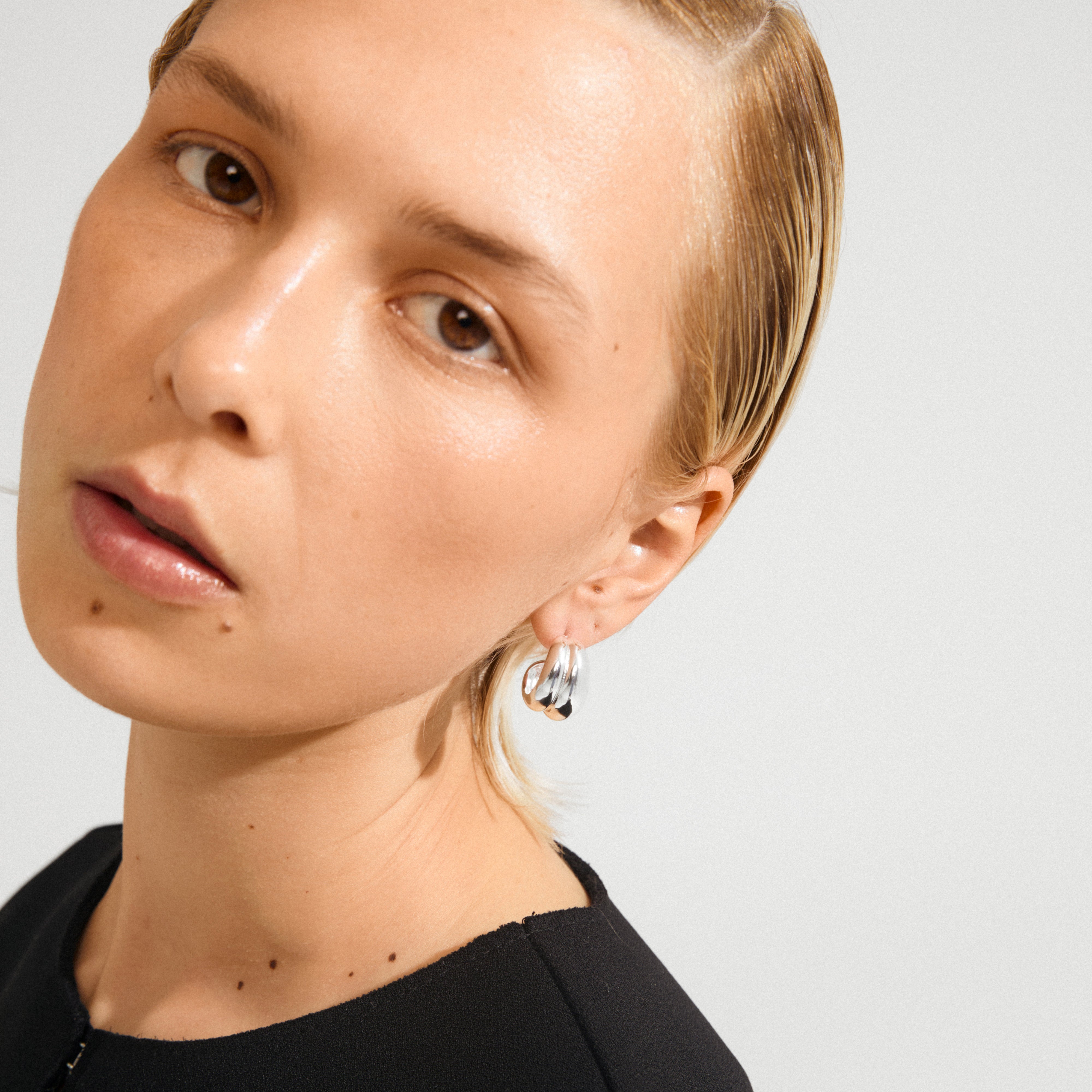 Young person wearing MIKO earrings silver-plated by Pilgrim, showcasing chunky, sculptural silver hoops against slicked-back hair and a black top.