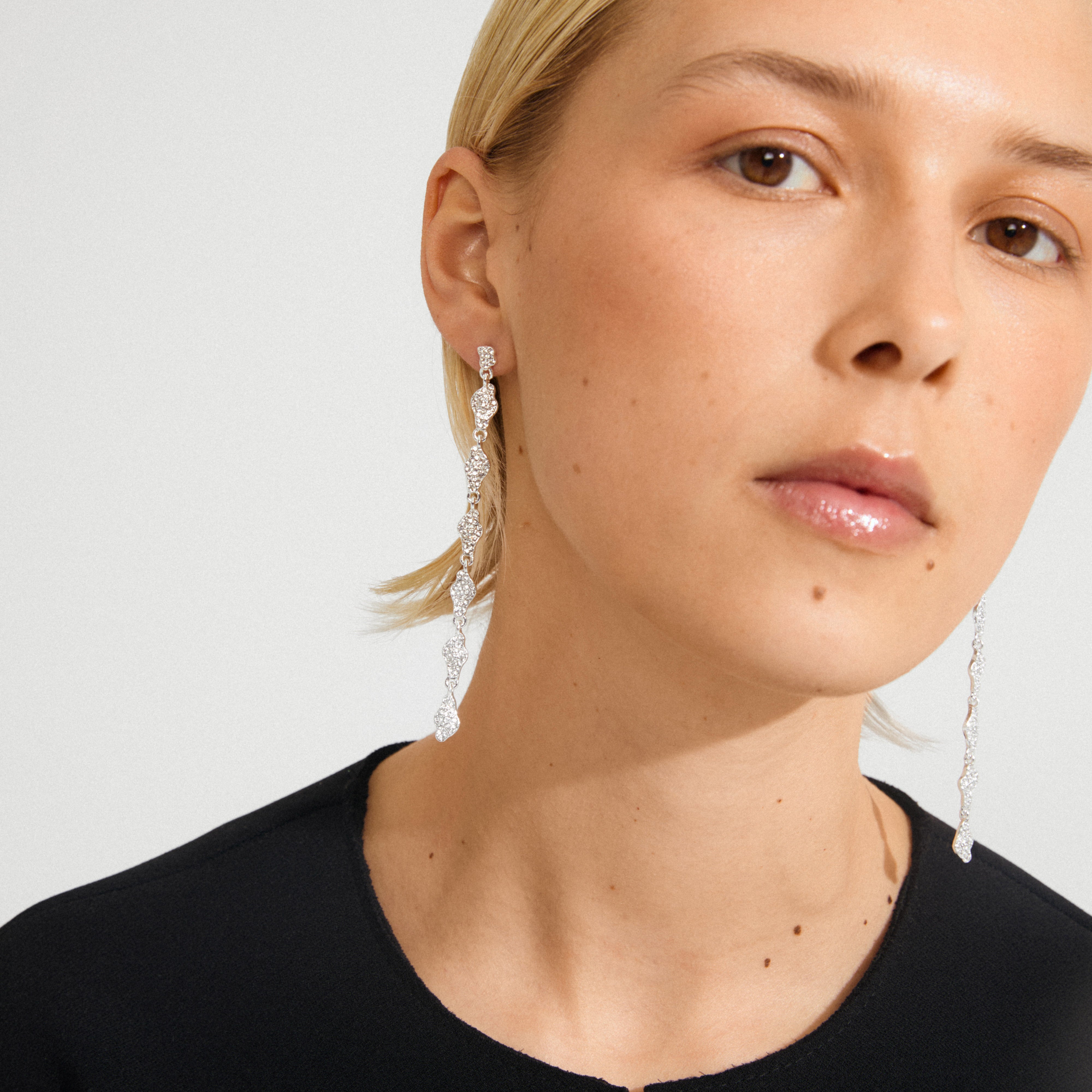 BASIL earrings silver-plated, featuring linked sparkle-encrusted elements, elegantly dangle from a young woman's ear, embodying Pilgrim's chic, minimalist aesthetic.