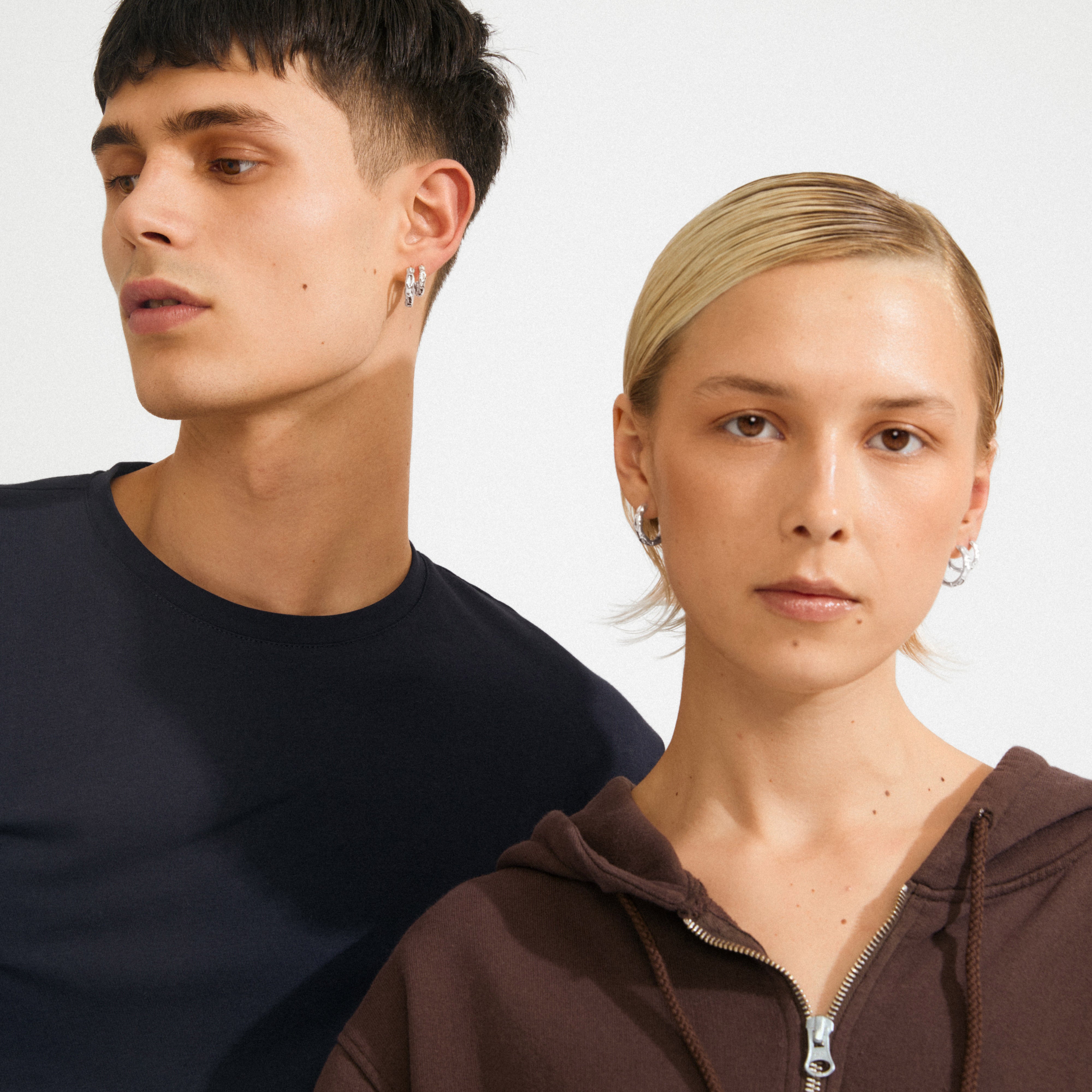 Two young adults showcase Pilgrim's DARBY silver-plated hoop earrings; the woman in a brown hoodie highlights their wavy design and versatile, stylish appeal.
