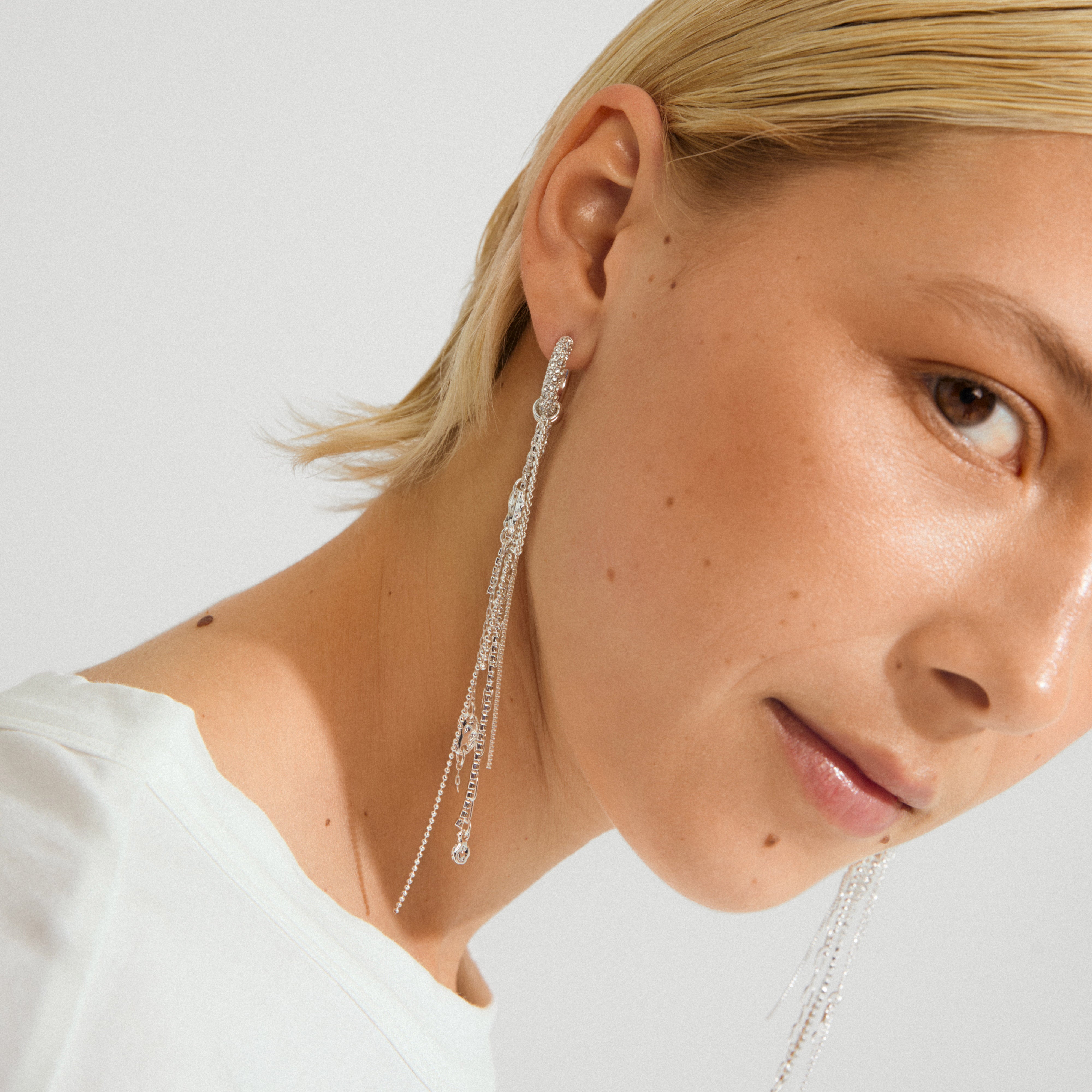 Young model wearing Pilgrim's DRUE earrings: silver-plated hoops with Preciosa crystals, cascading chains, and pendants, showcasing a luxurious bohemian style.