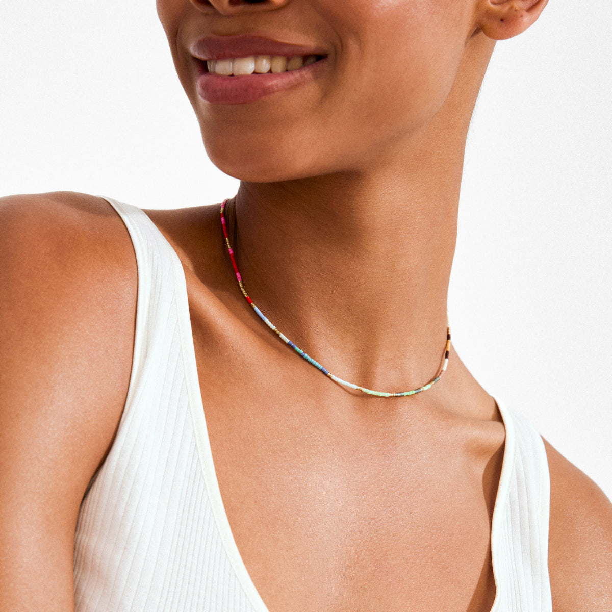 Person wearing ALISON necklace multi, gold-plated with colorful beads, paired with a white rib-knit tank top, showcasing a playful and vibrant style.