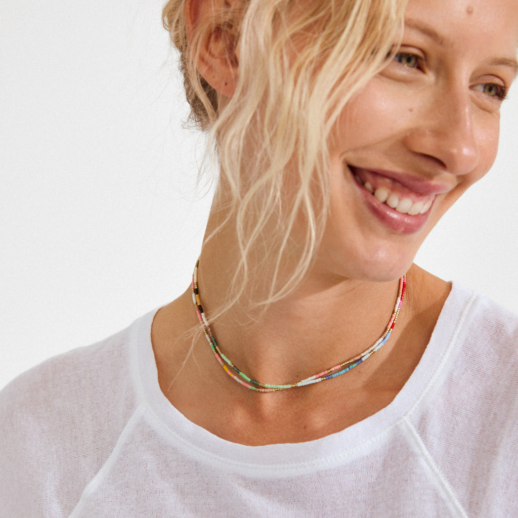 Smiling woman wearing Pilgrim's ALISON necklace, a double-strand, multicolored beaded piece, paired with a simple white top for a vibrant, carefree look.