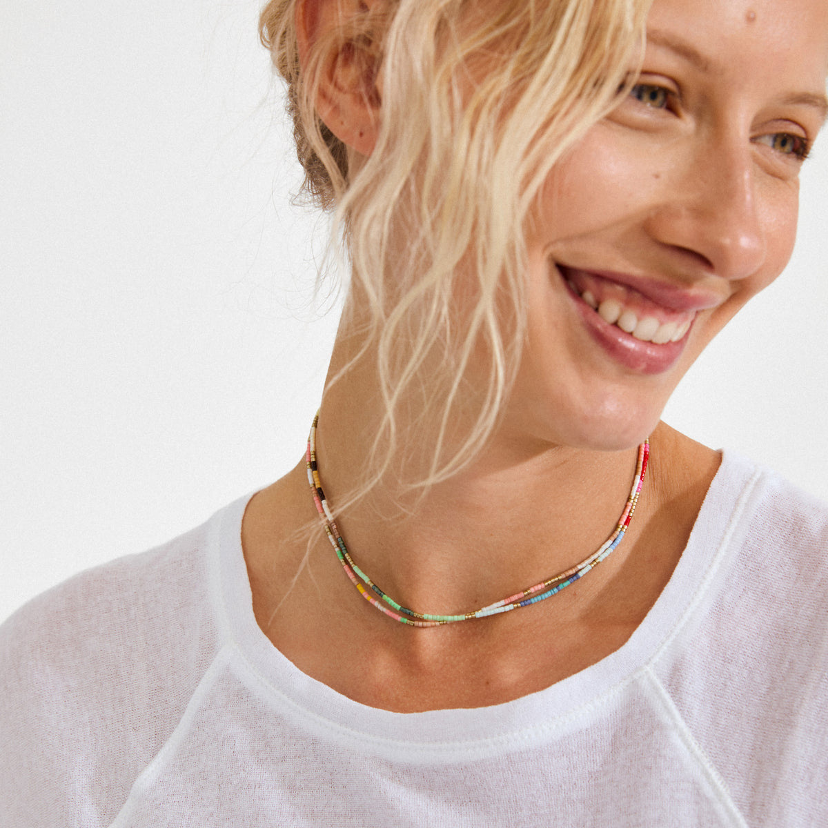 Smiling woman wearing Pilgrim's ALISON necklace, a double-strand, multicolored beaded piece, paired with a simple white top for a vibrant, carefree look.