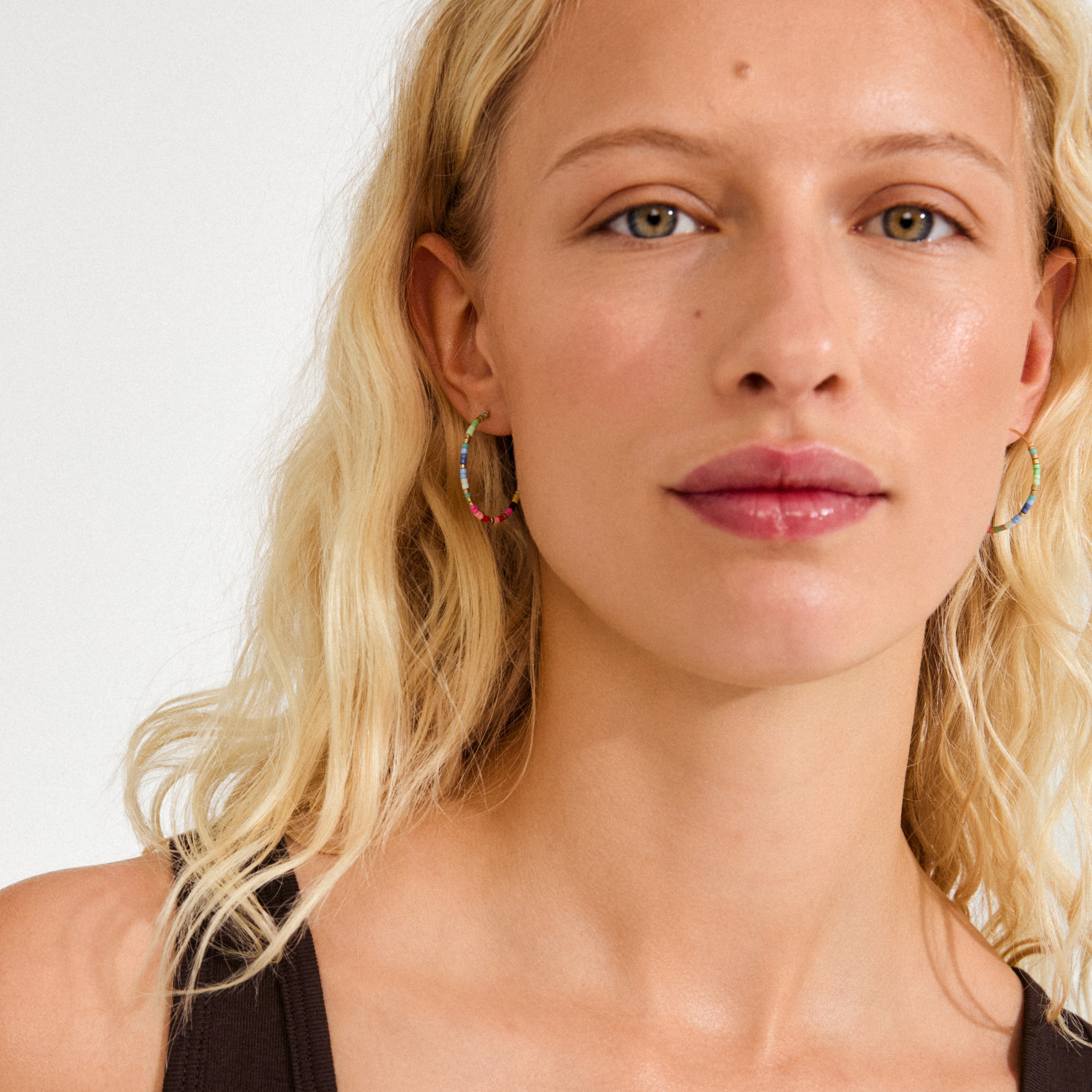 Young woman wears Pilgrim's ALISON earrings, gold-plated hoops adorned with colorful beads, complementing her wavy blonde hair and black tank top.