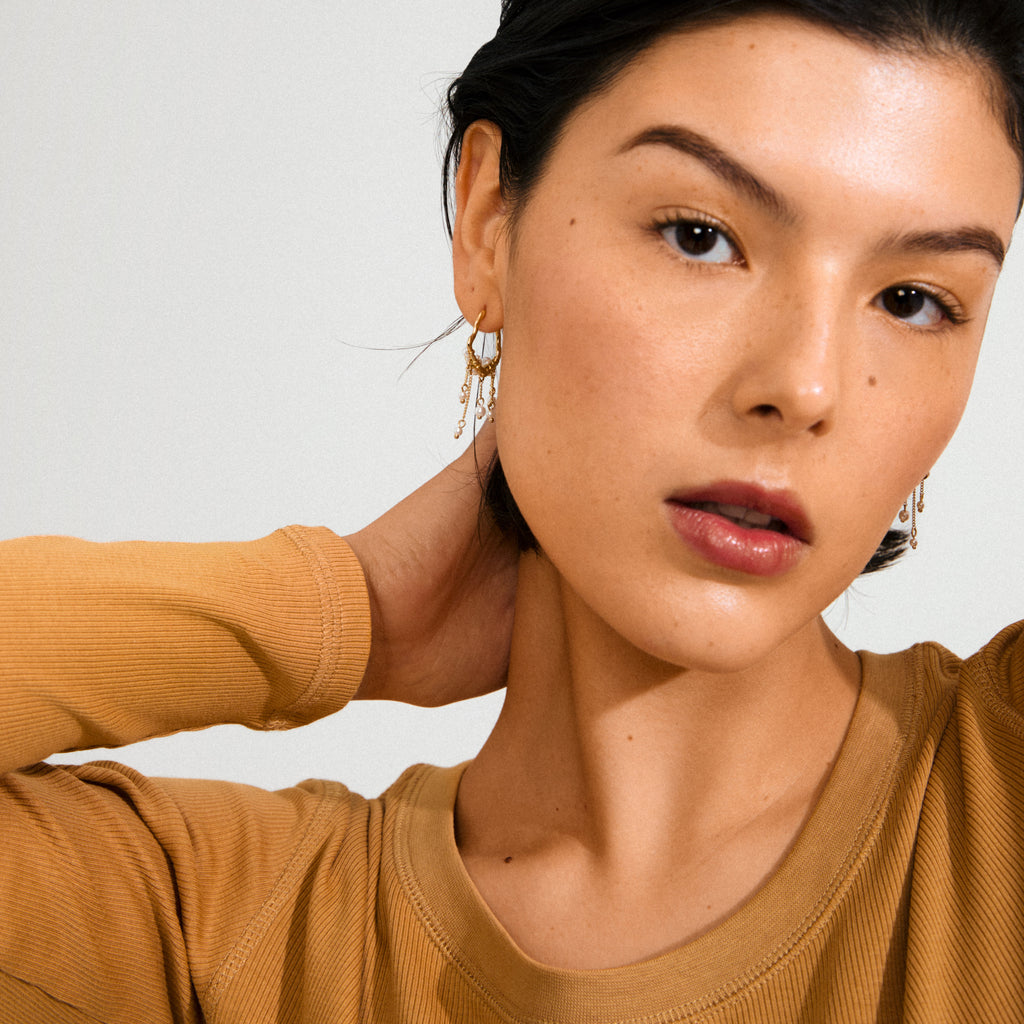 JASPER earrings gold-plated feature dreamcatcher-inspired design with pearls and delicate chains, worn by a woman in a tan ribbed top and gold hoops.
