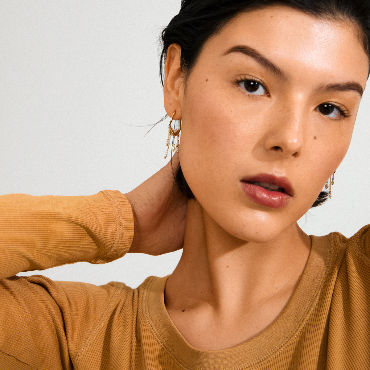 JASPER earrings gold-plated feature dreamcatcher-inspired design with pearls and delicate chains, worn by a woman in a tan ribbed top and gold hoops.