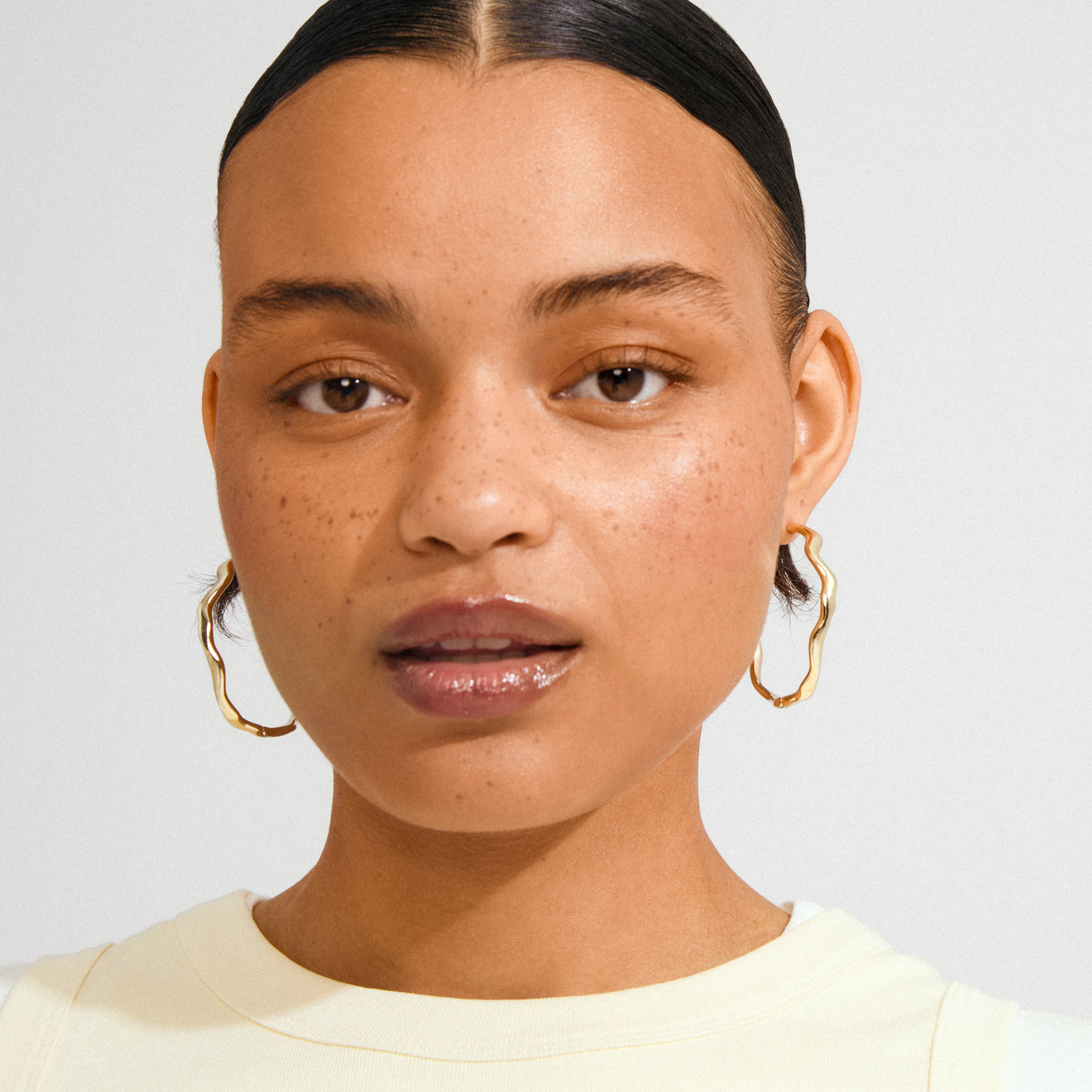 Young woman wearing Pilgrim's LARKIN gold-plated earrings, featuring wavy, textured hoops that catch light, paired with a pale yellow top.