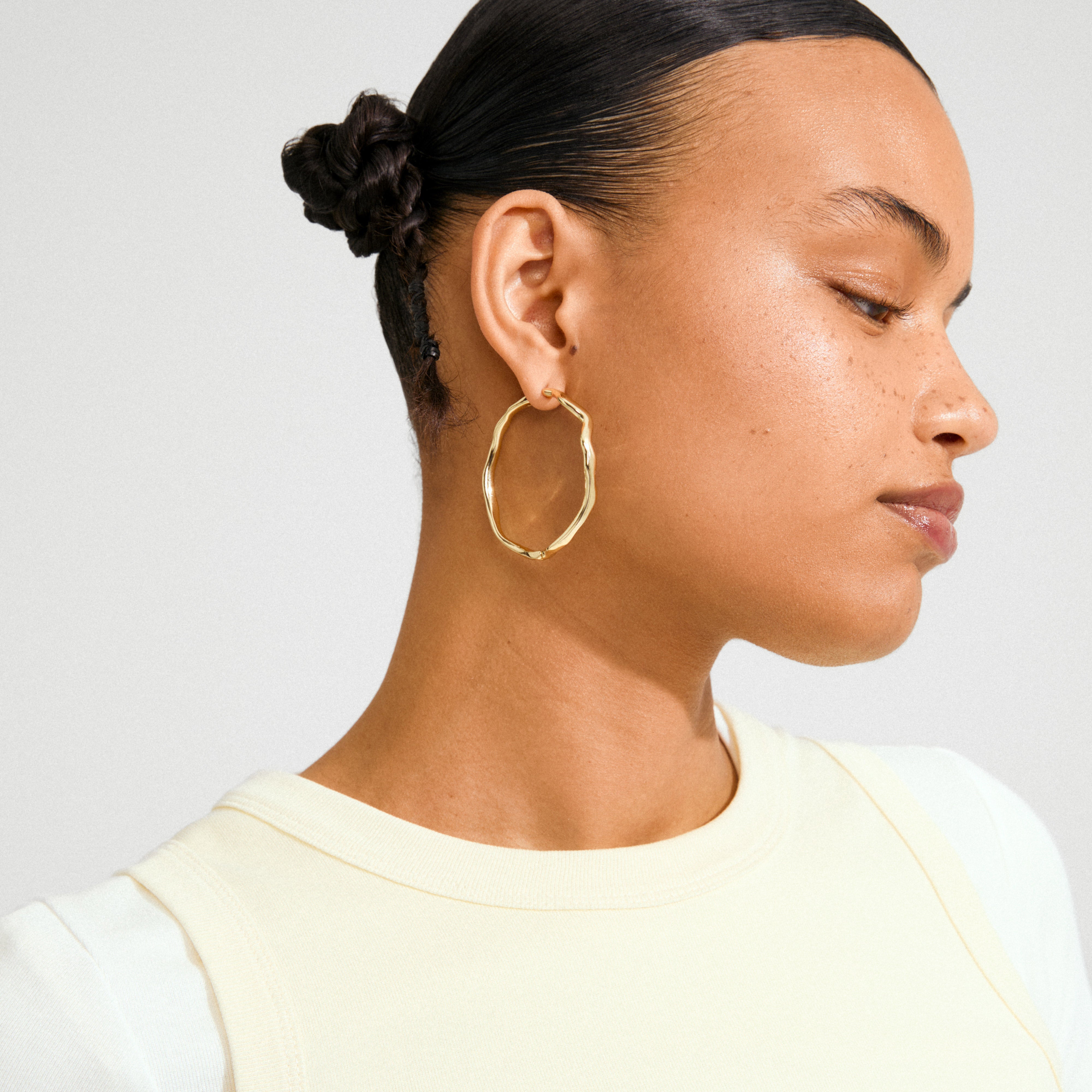 Woman wearing Pilgrim's LARKIN gold-plated earrings, featuring a wavy, textured hoop design, highlighting minimalist elegance and light-catching detail. Hair in braided bun, showcasing earring's impact.