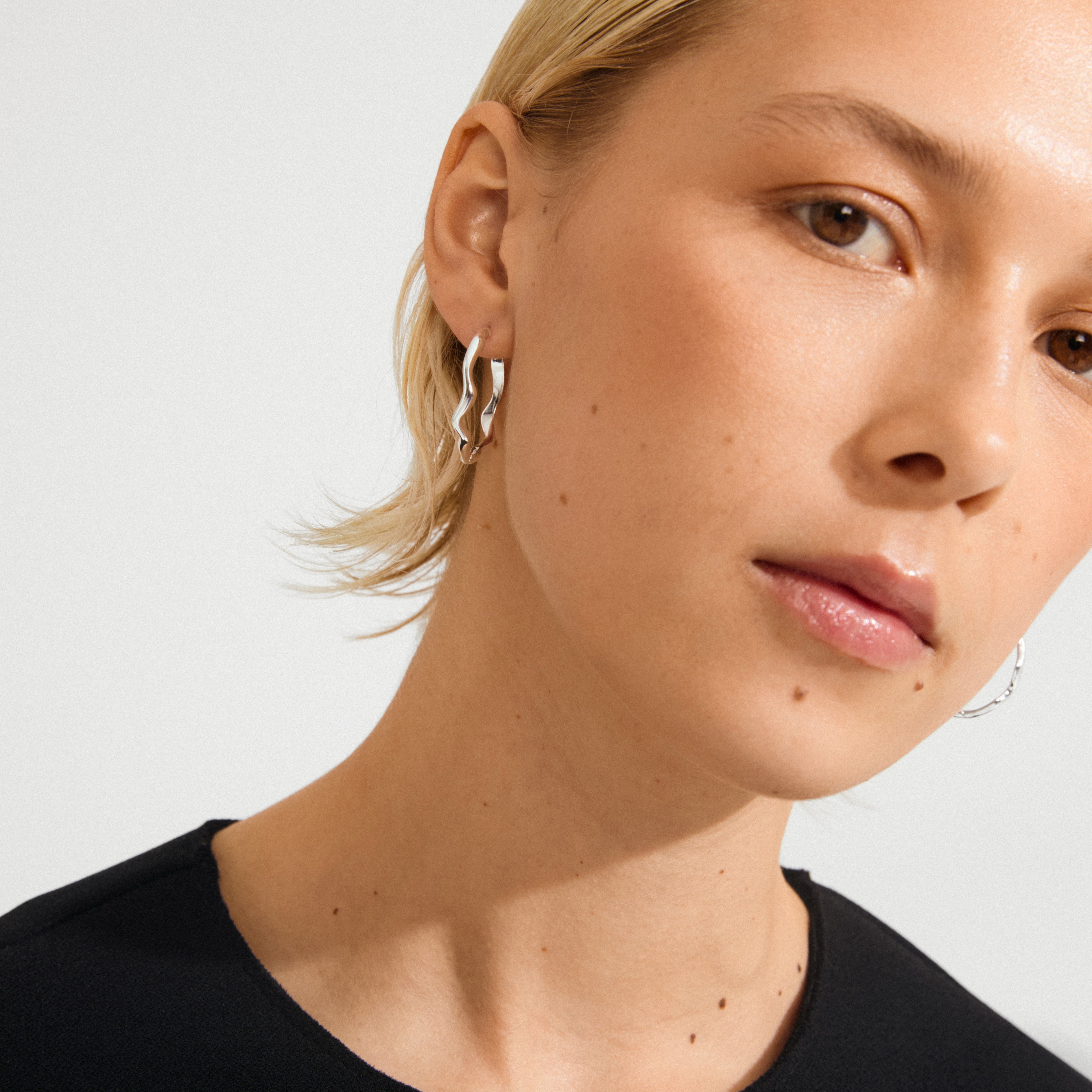 Young woman wears Pilgrim's LARKIN silver-plated earrings, featuring wavy, nature-inspired hoops that catch light, highlighting minimalist design against her black top.