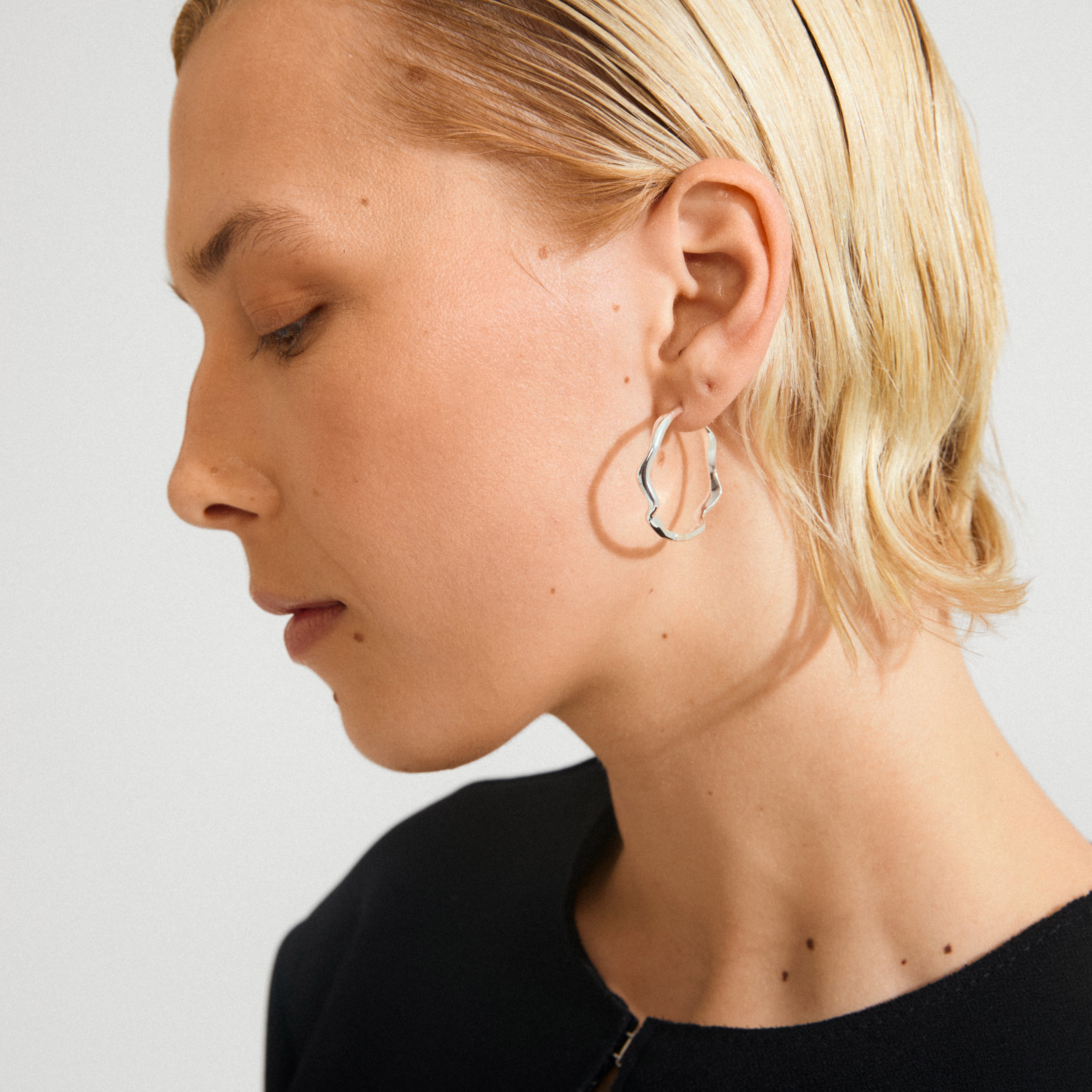 Close-up of a person wearing LARKIN silver-plated wavy hoop earrings, showcasing their minimalist design and sleek silhouette against a light backdrop.