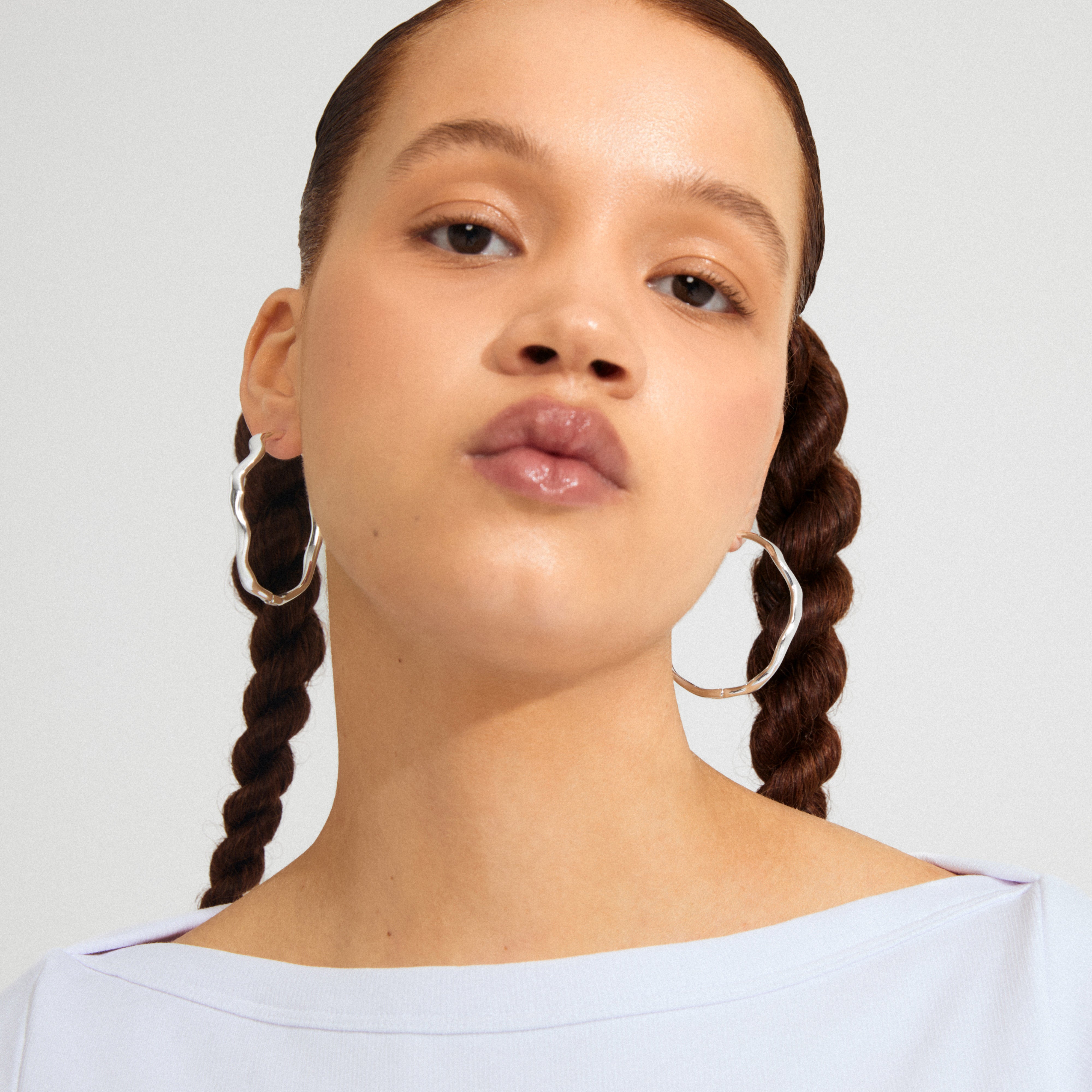 Young person wearing large, wavy LARKIN earrings, silver-plated, with natural makeup and braided hair, showcasing Pilgrim's stylish, minimalist design.