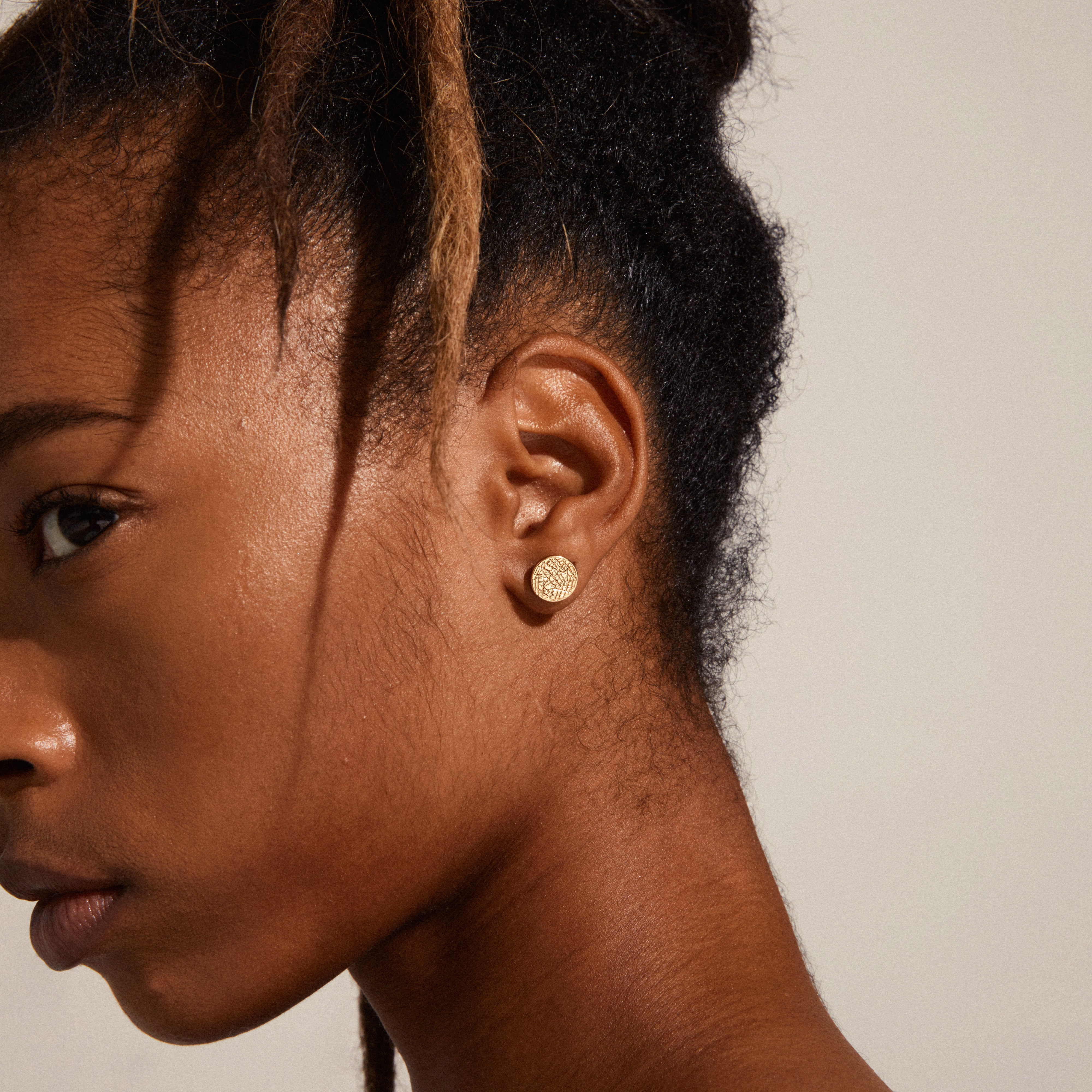 Gold-plated WYNONNA rustic disc earrings with a raw pattern, showcased on a woman's ear, offering a blend of edgy and elegant style.