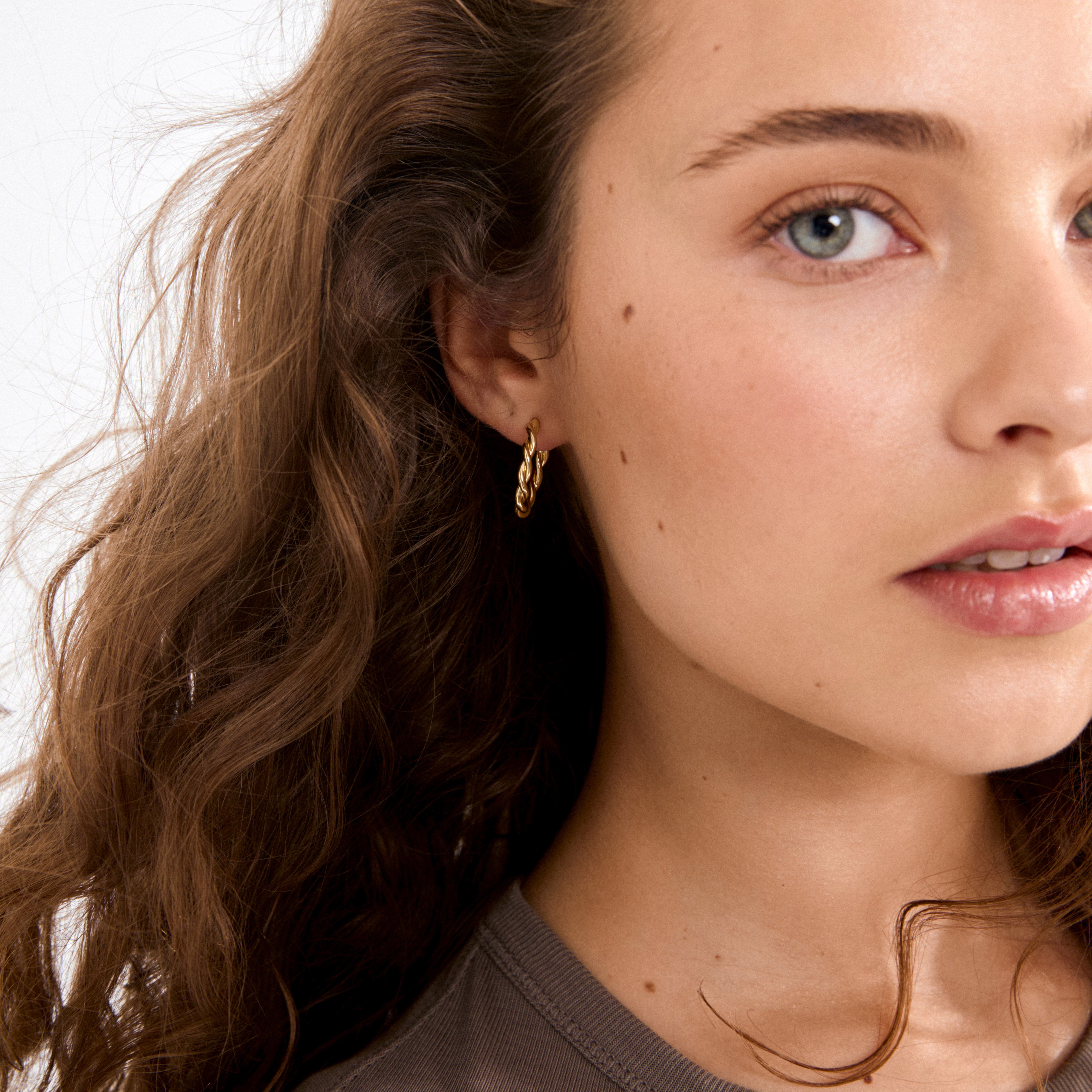 Young woman wearing NAJA twisted hoop earrings, gold-plated, showcasing their sculptural, light-catching design against curly brown hair and a neutral-toned top.