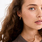 Young woman wearing NAJA twisted hoop earrings, gold-plated, showcasing their sculptural, light-catching design against curly brown hair and a neutral-toned top.