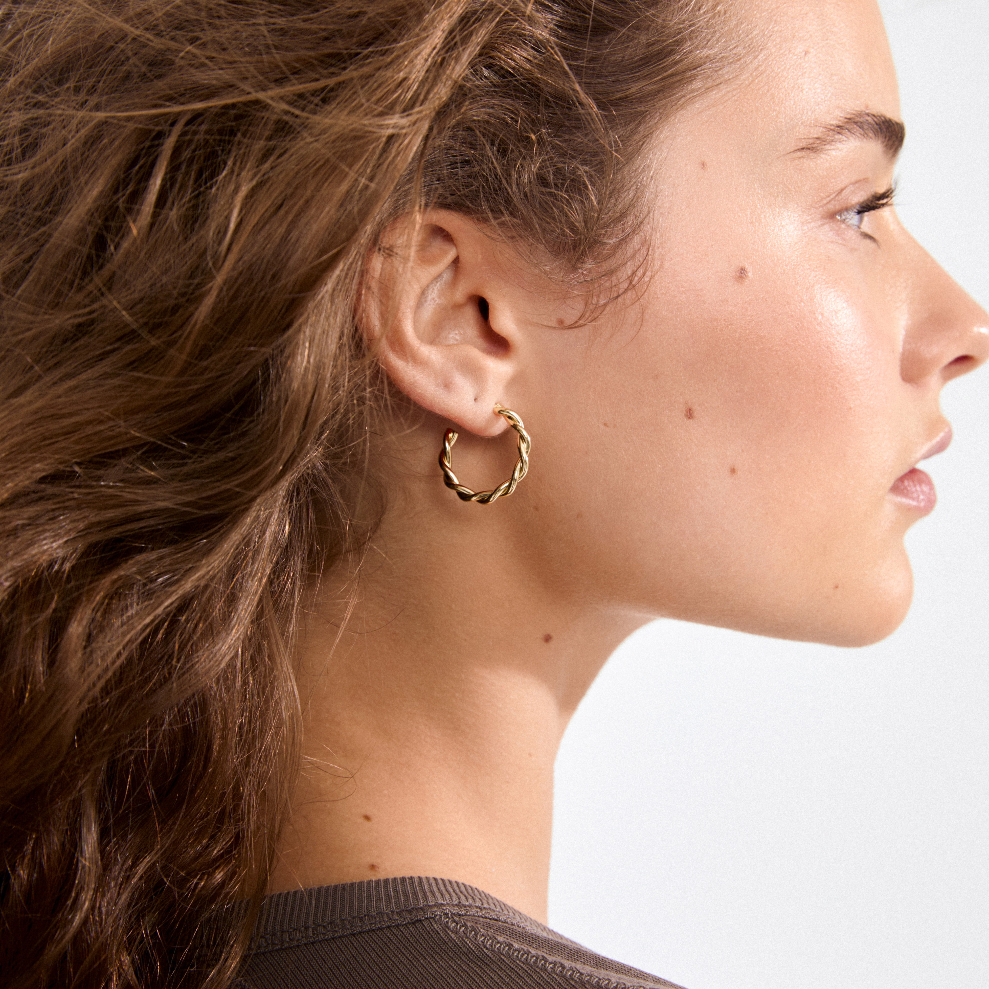 Close-up of a young woman wearing NAJA twisted hoop earrings, gold-plated. The sculptural design catches light, highlighting her smooth skin and subtle makeup.