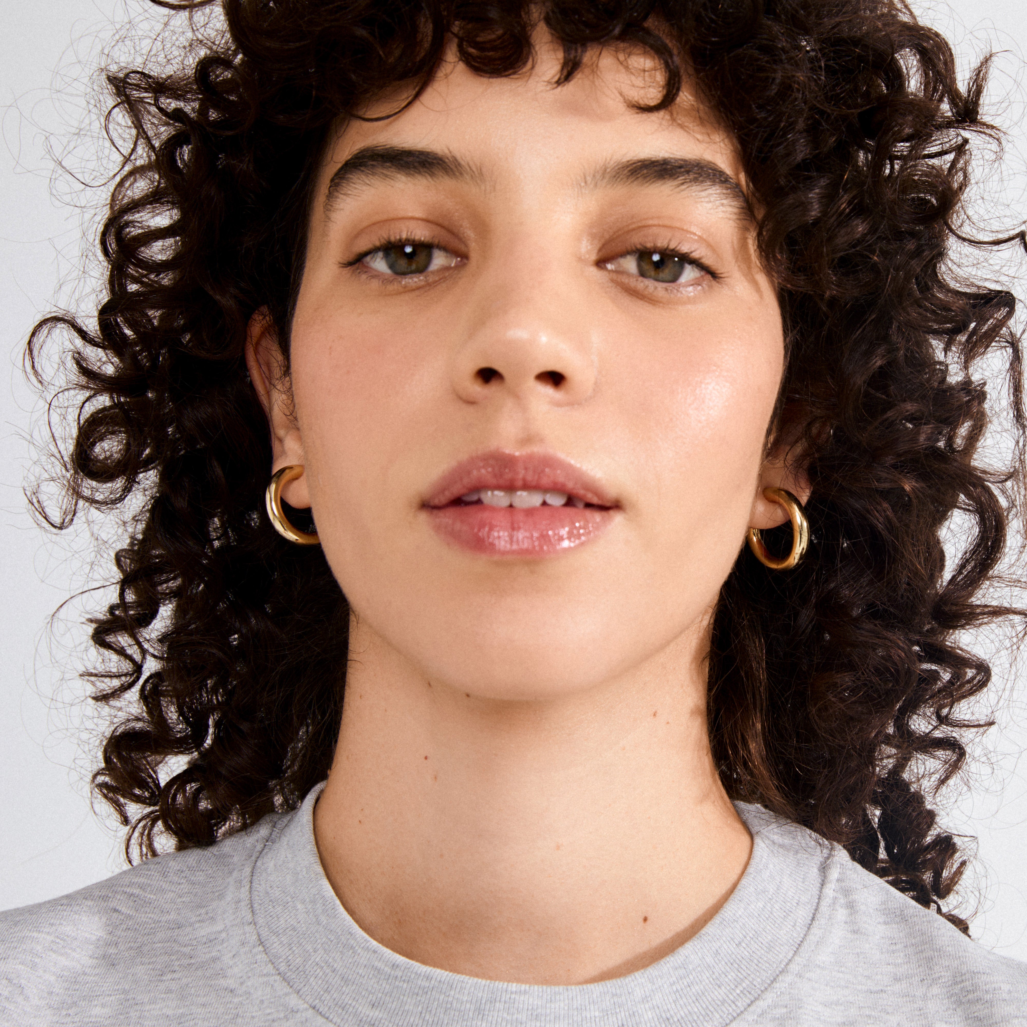 Young adult wearing MADDIE chunky hoop earrings, gold-plated, with a glossy finish, highlighting their bold, stylish appeal against a simple gray tee.