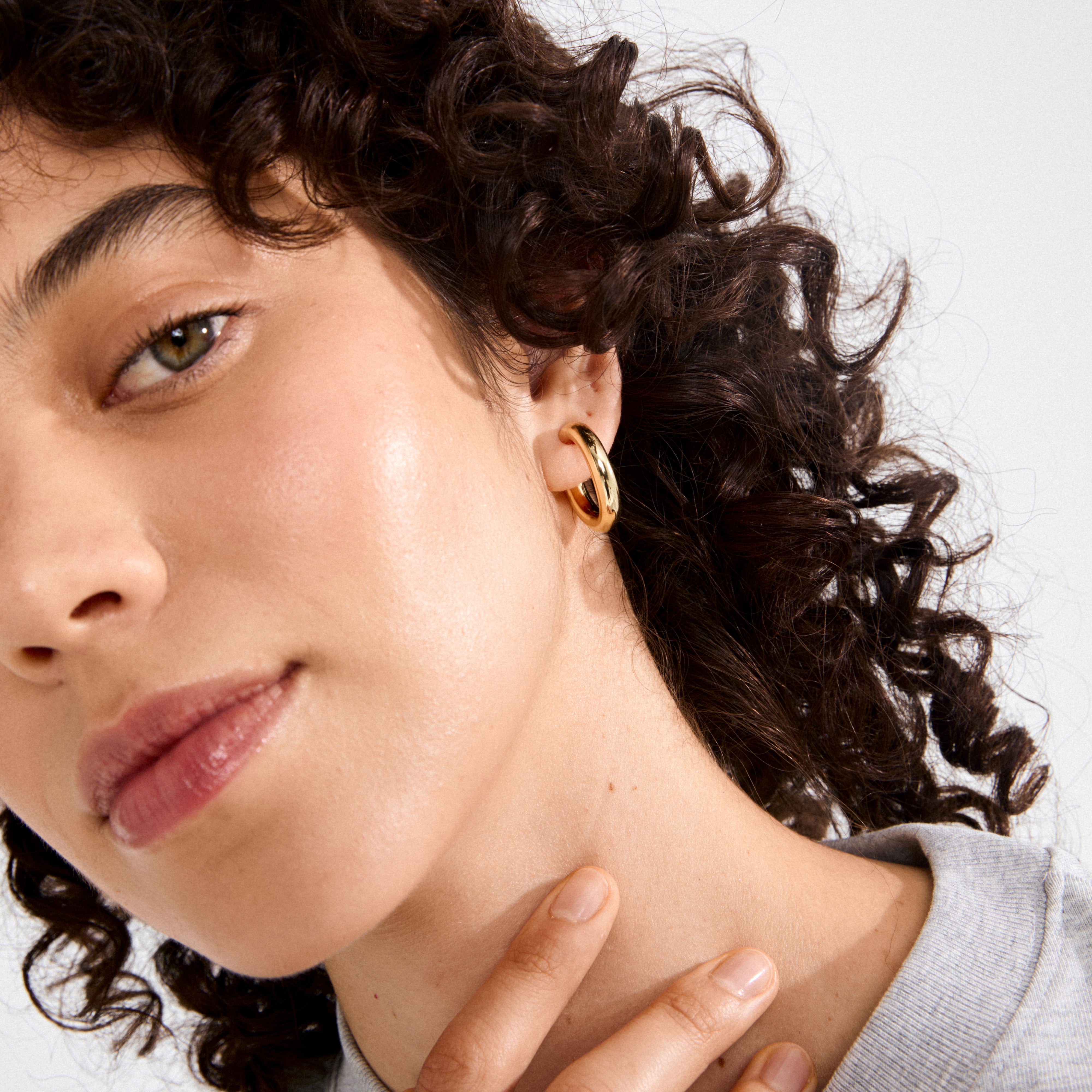 Close-up of a face showcasing MADDIE chunky hoop earrings, gold-plated, with a glossy finish, worn in the left ear amidst curly hair.