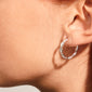 NAJA twisted hoop earrings in shiny silver-plated metal, featuring a twisted-rope design that elegantly catches light, worn on an ear, showcasing Pilgrim's Scandinavian craftsmanship.