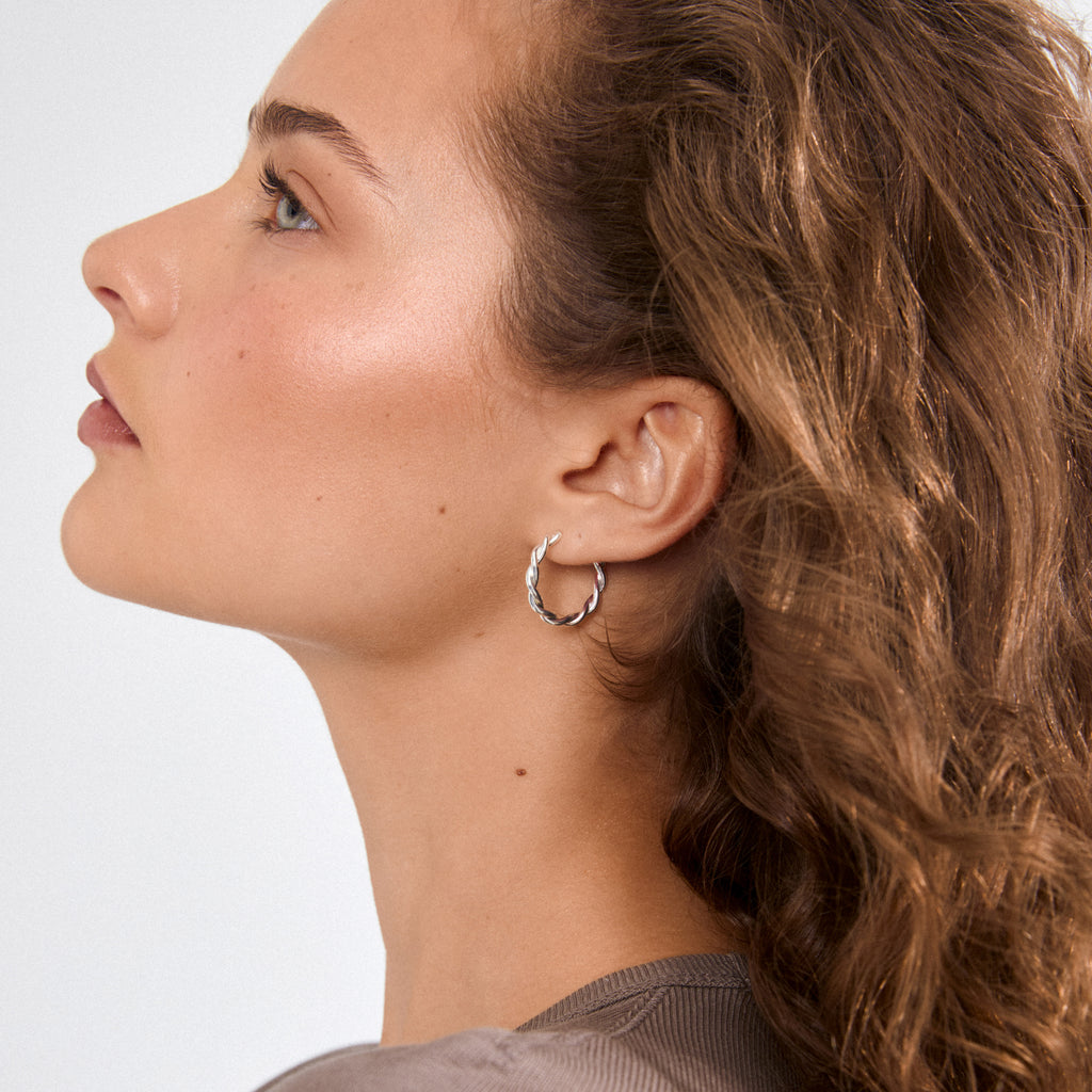 Close-up of NAJA twisted hoop earrings, silver-plated and sculptural, worn by a person with long brown hair, showcasing the hoop's stylish, playful design.
