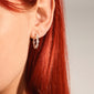 Close-up of a left ear wearing NAJA twisted hoop earrings, silver-plated, showcasing their elegant, light-catching design amidst bright red hair.