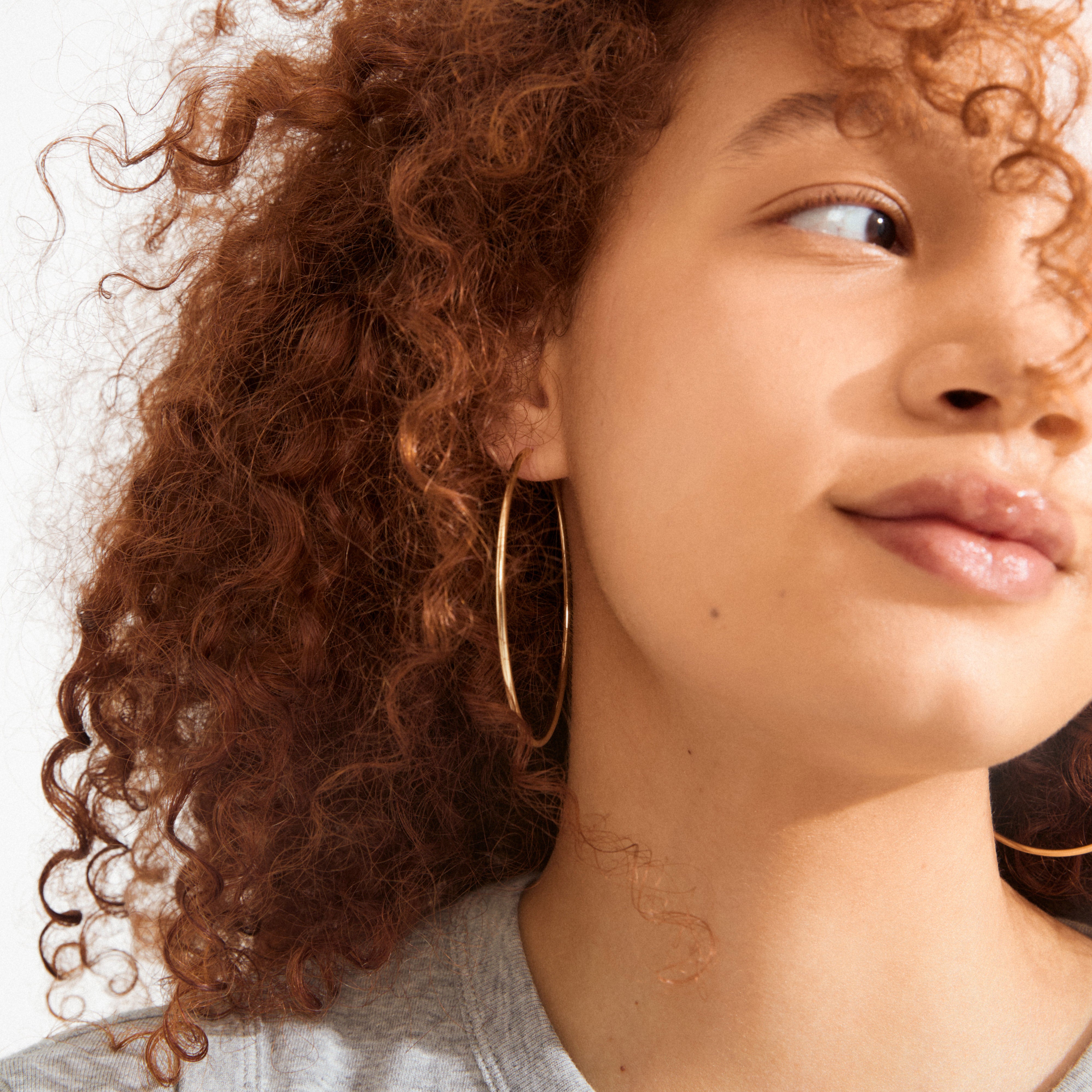 Young person wearing APRIL large gold-plated hoop earrings, 60 mm in diameter, with a slim, stylish design. Earrings complement their curly hair and light top.